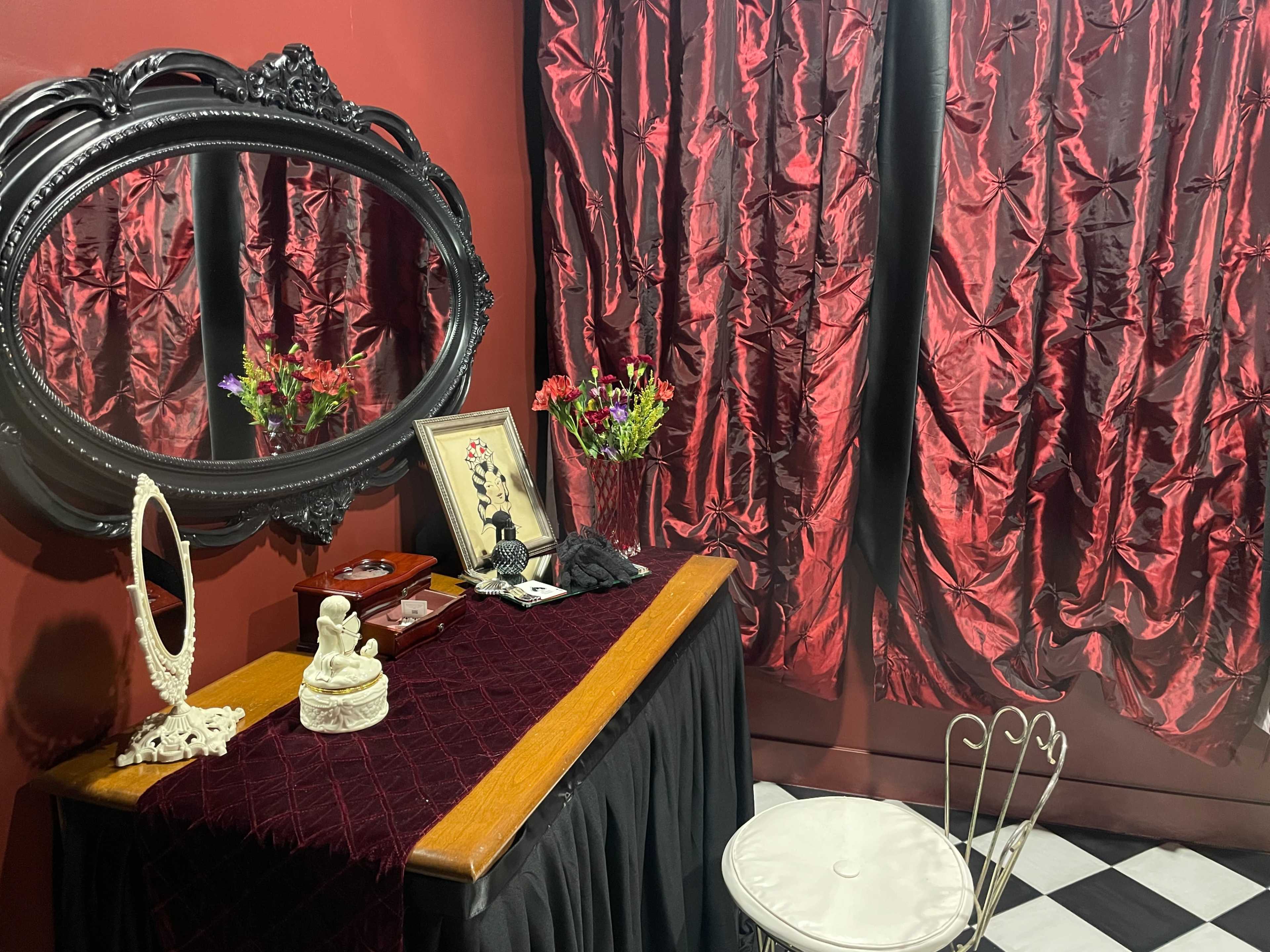The image features a vintage vanity table with a mirror, surrounded by red velvet curtains and a decorative chair on a black and white checkered floor.