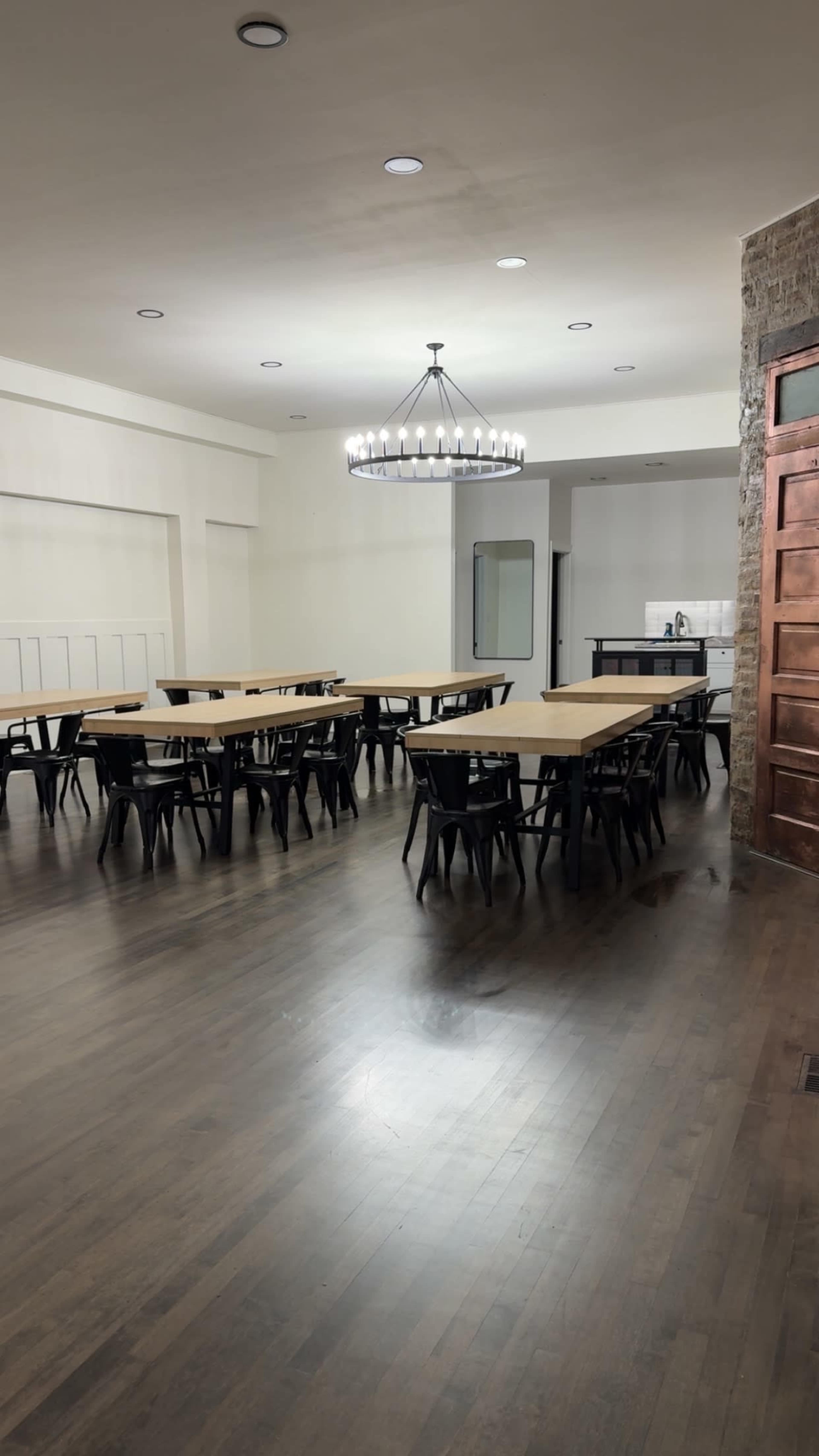 The image shows a spacious room with multiple rectangular tables and black chairs arranged neatly, a chandelier overhead, and a small kitchen area visible in the background.