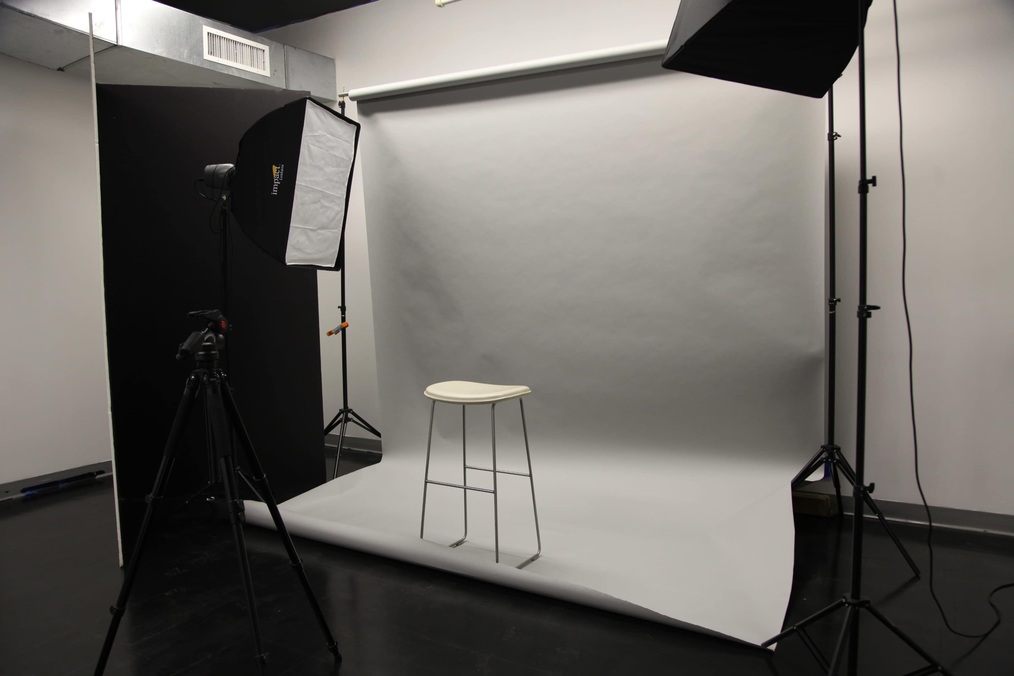 The image shows a photography studio setup with a gray backdrop, a stool in the center, and lighting equipment positioned nearby.