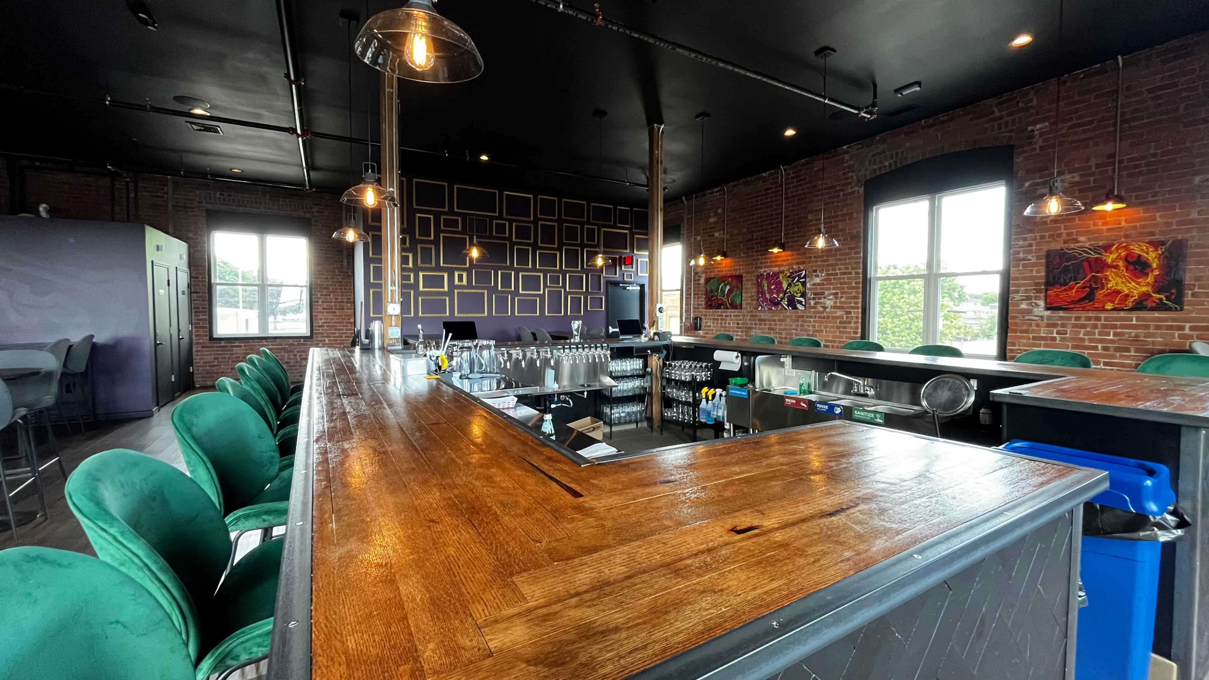 A stylish bar area with a wooden counter, green upholstered stools, and large windows revealing a brick wall adorned with framed artwork.