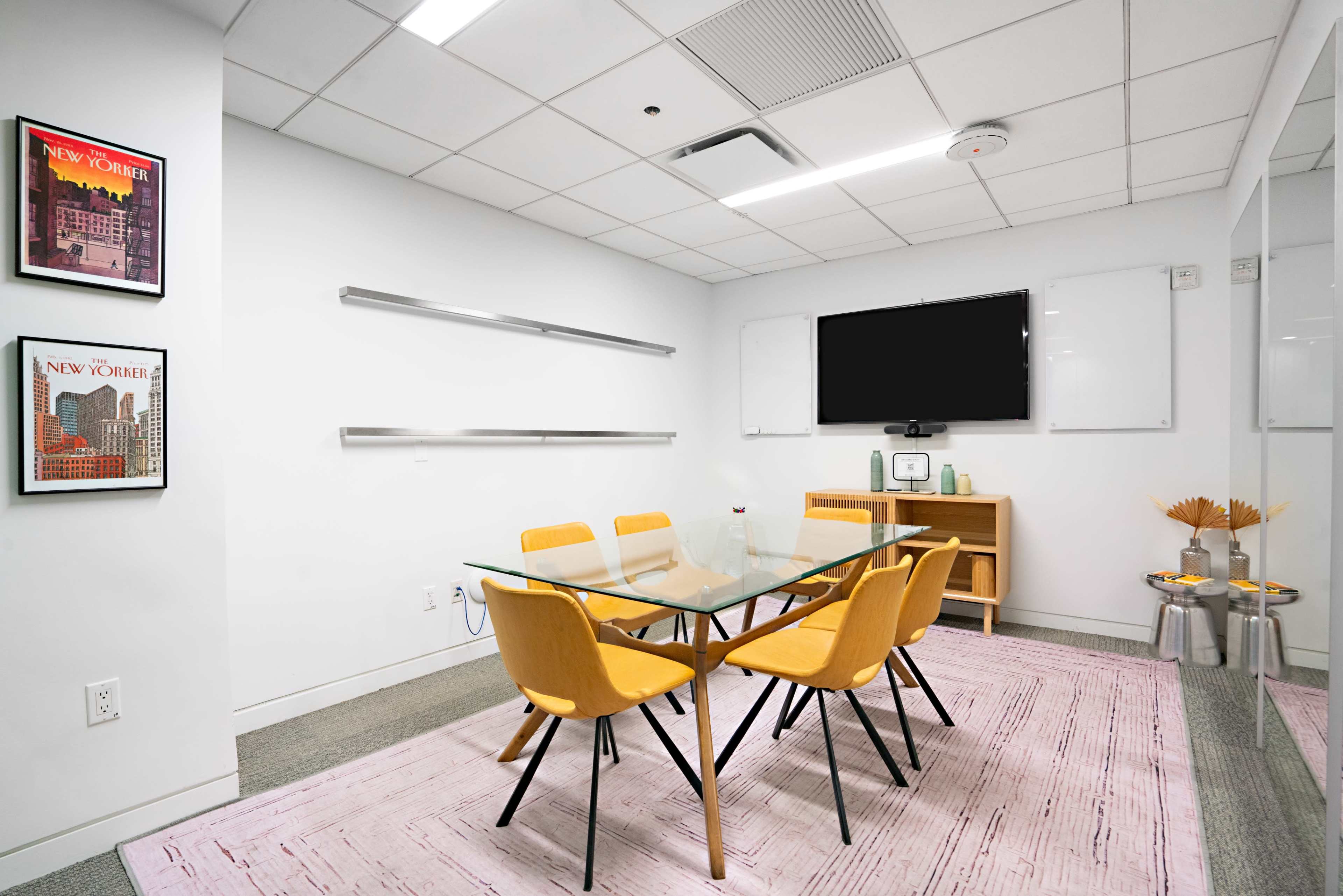 A modern meeting room features a glass table surrounded by yellow chairs, with a large screen on the wall and various wall-mounted fixtures.