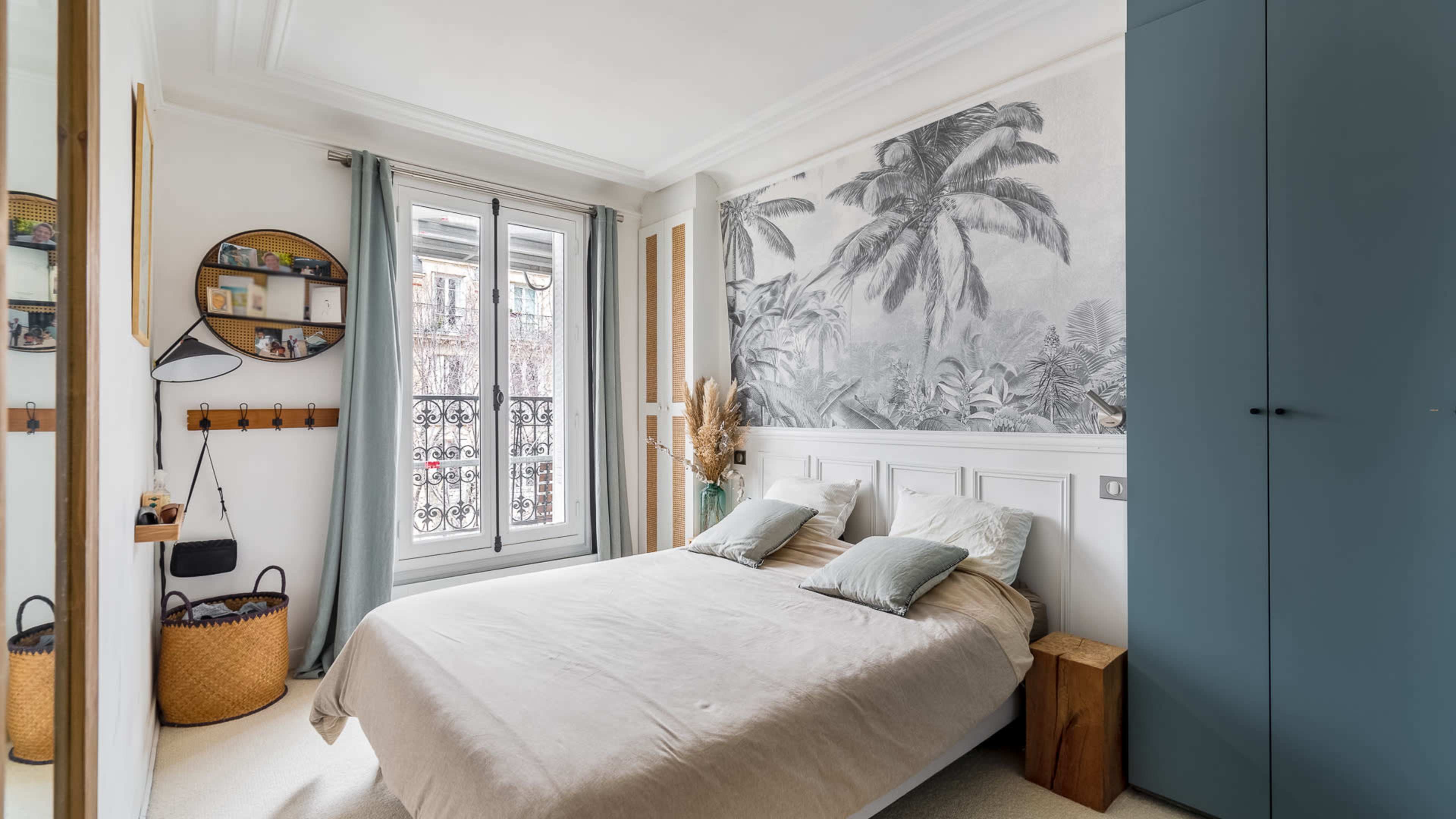A light-colored bedroom featuring a bed with two pillows, a large mural of palm leaves on the wall, and a window with sheer curtains.