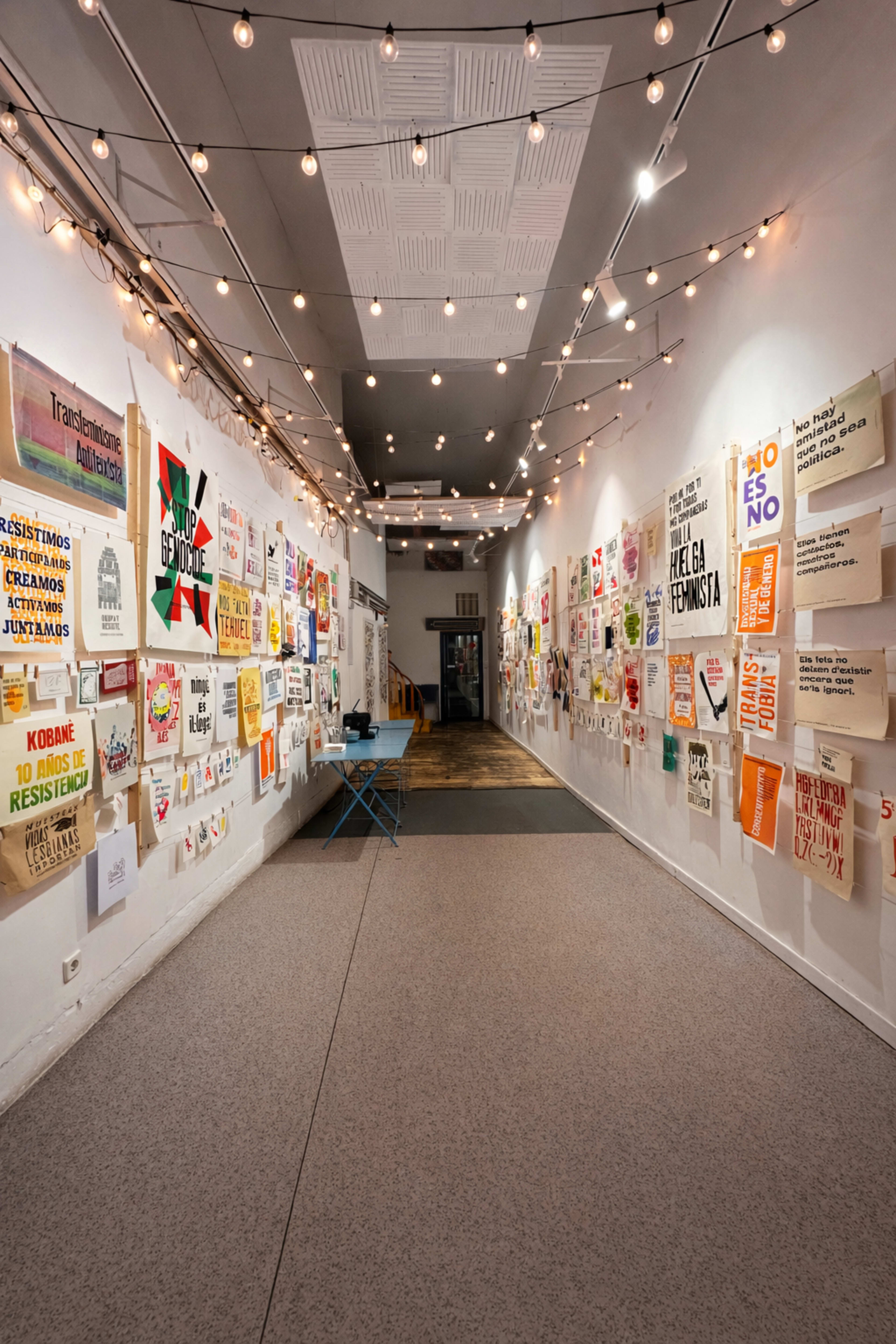 The image shows a narrow hallway illuminated by string lights, lined with colorful posters on the walls.