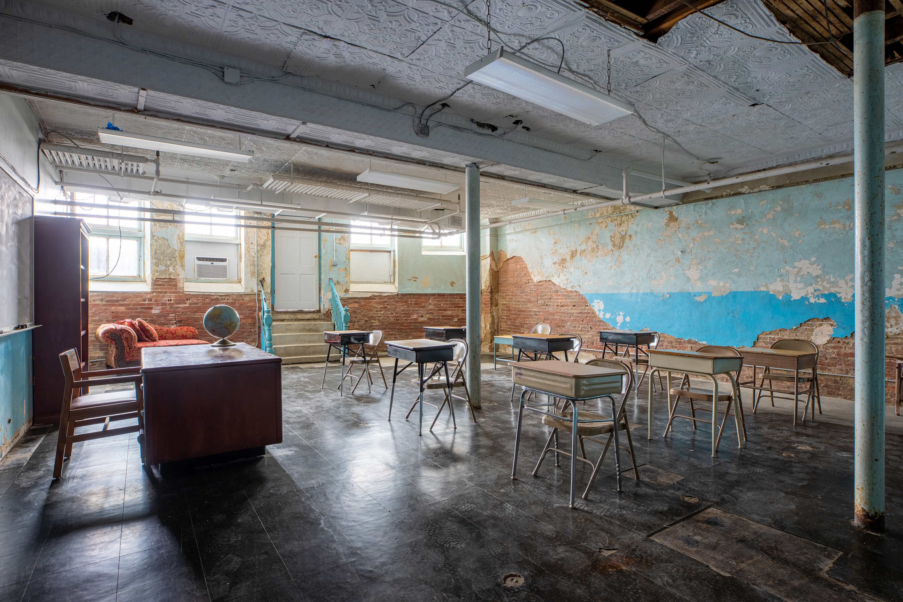 An empty classroom features a wooden desk and several metal tables arranged around peeling blue walls and a partially exposed brick structure.