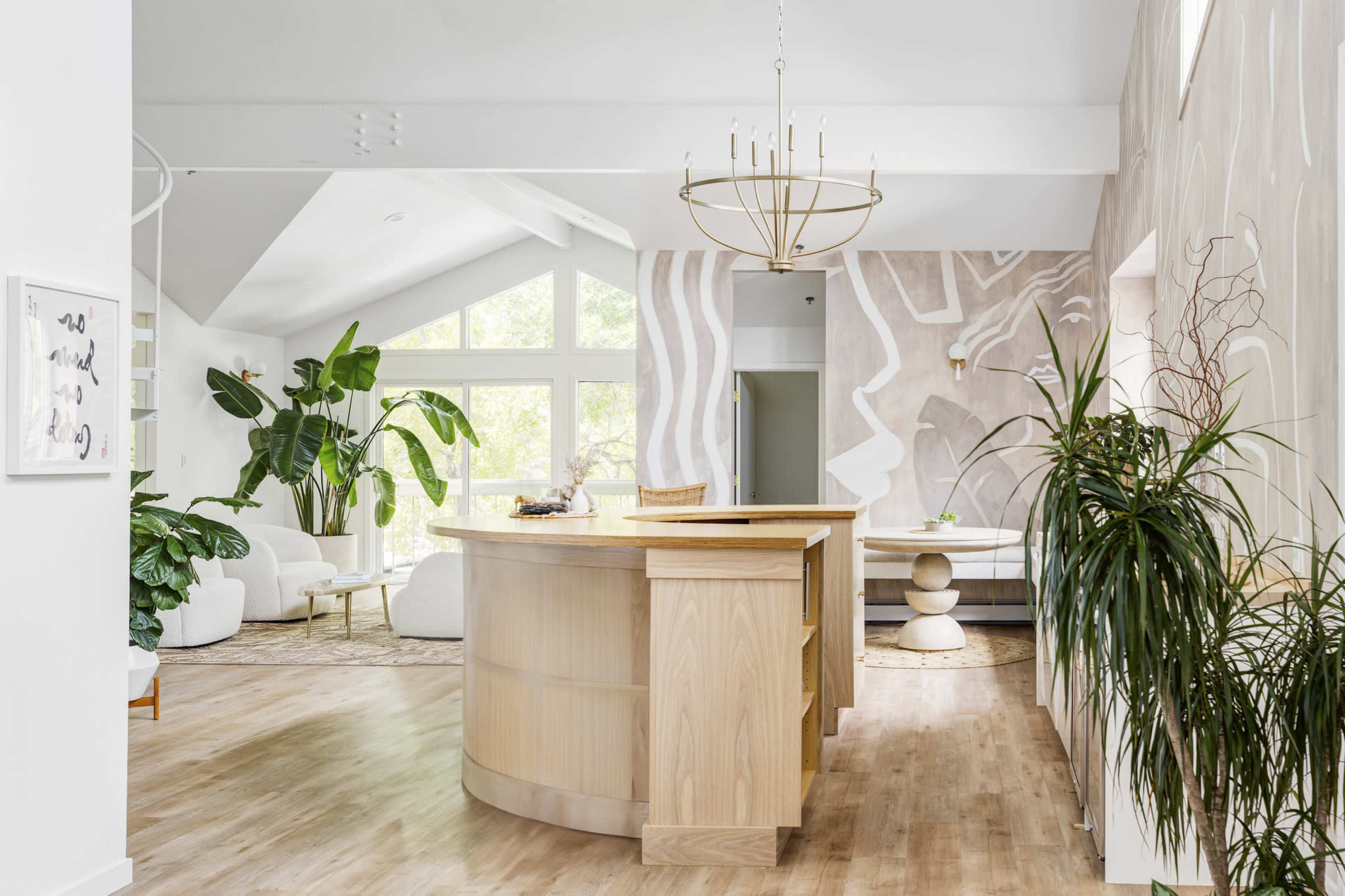 The image depicts a modern, open-concept interior featuring a light wooden reception desk, large windows filled with natural light, and lush green plants surrounding the space.