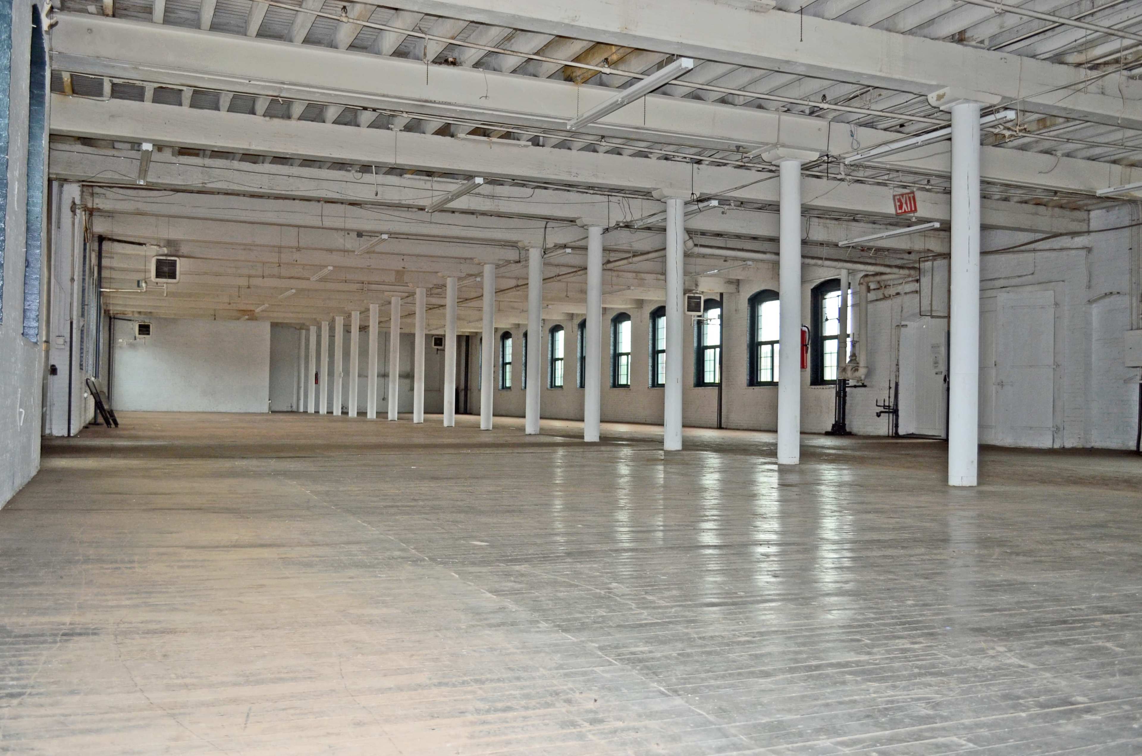 An empty industrial space features wooden floors, white walls, and multiple columns supporting the ceiling.