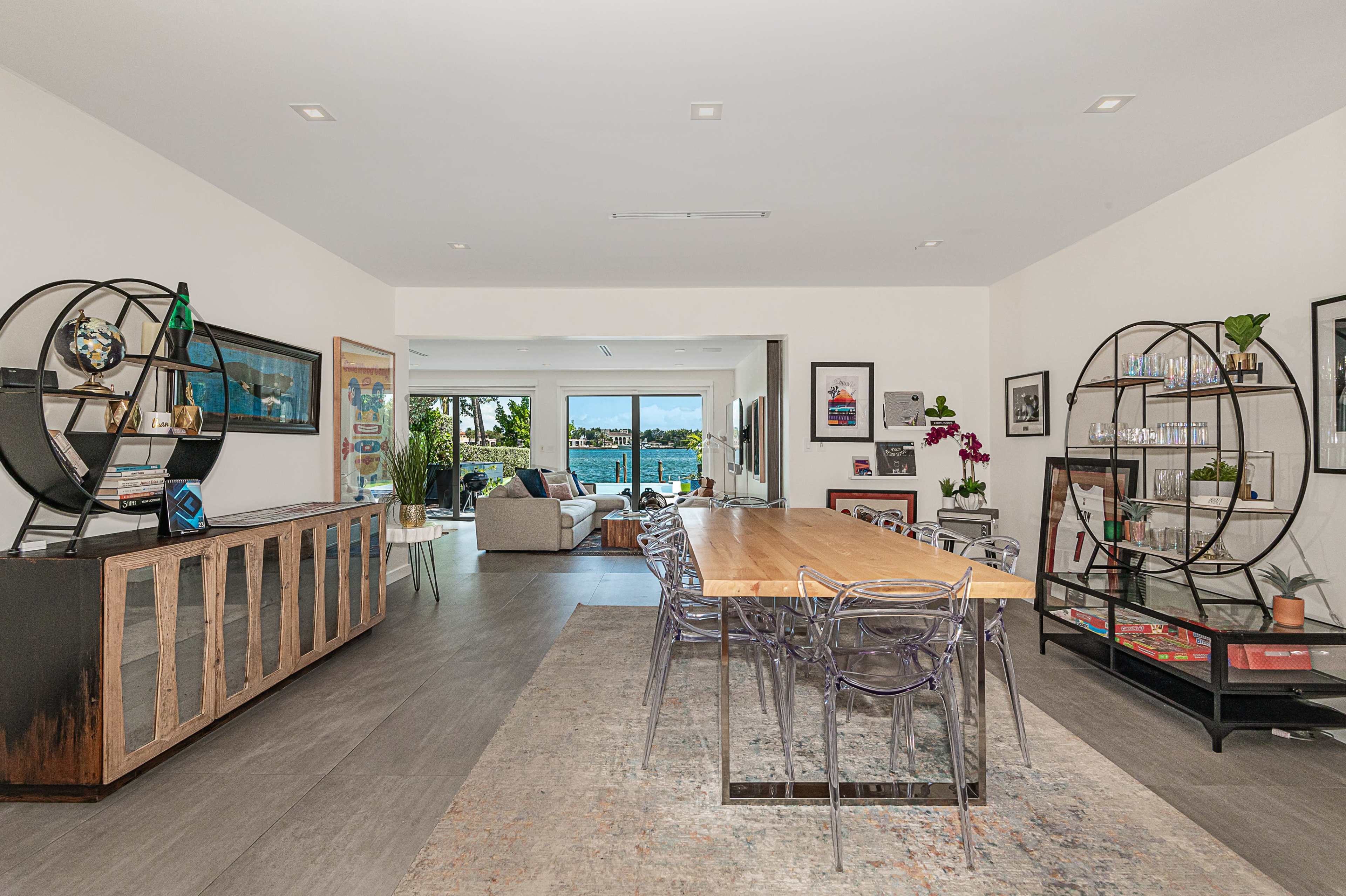 The image shows a modern living and dining area with a long wooden table, transparent chairs, and open views of a waterfront through large windows.