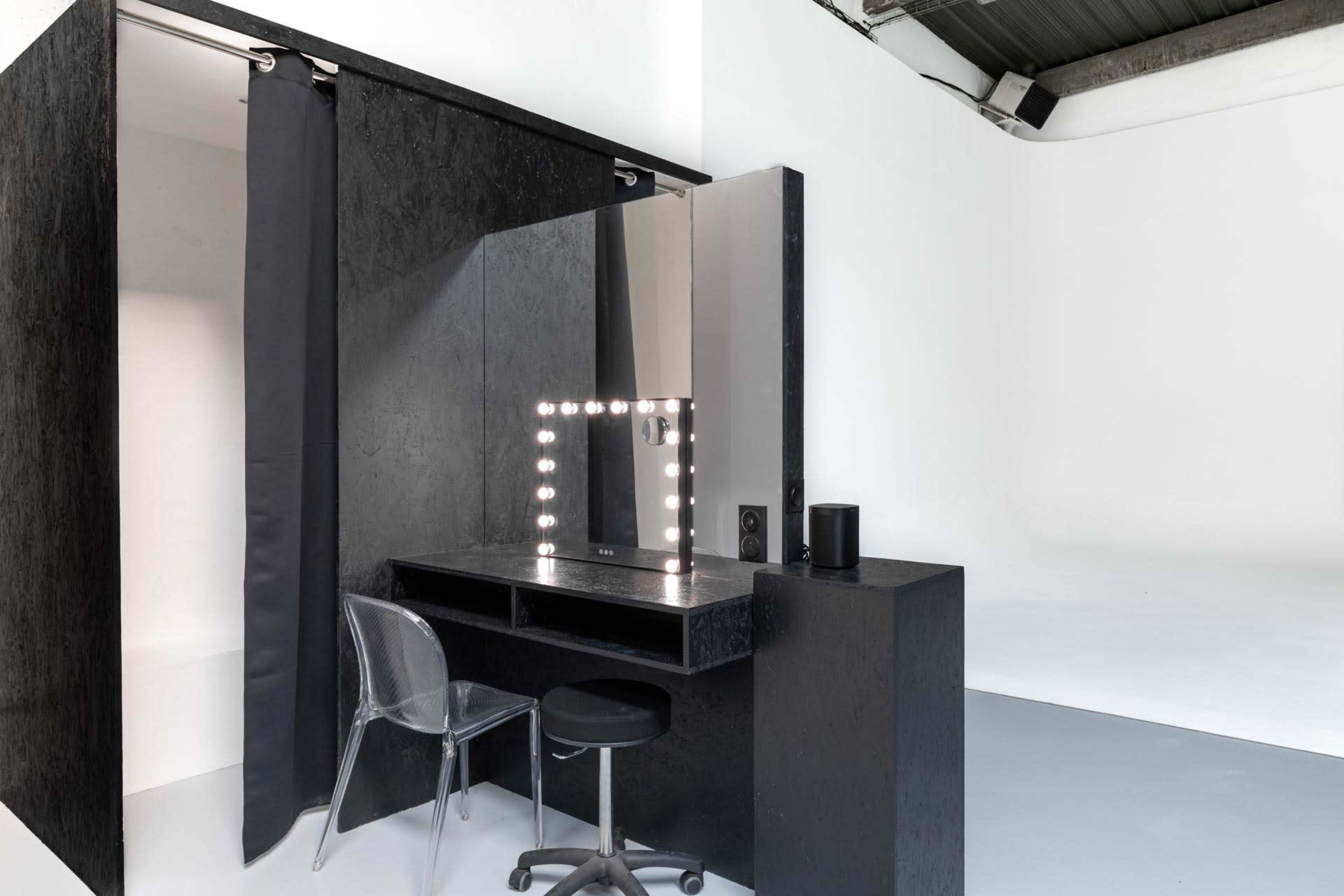 A minimalist makeup station featuring a mirror with lights, a black desk, a transparent chair, and a stool in a studio with a white backdrop.