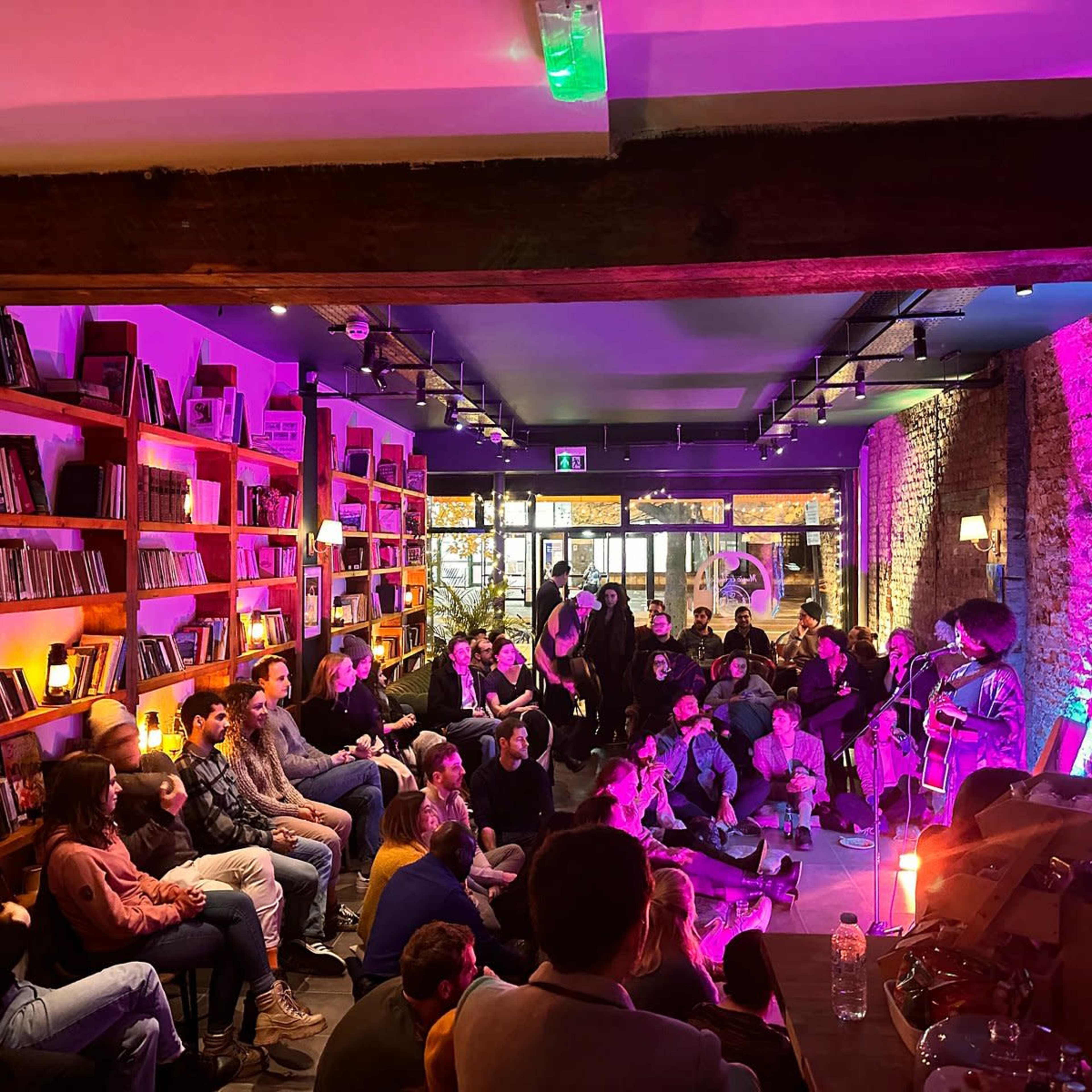 A musician performs on stage in a dimly lit venue filled with seated listeners, surrounded by shelves of records and soft purple lighting.
