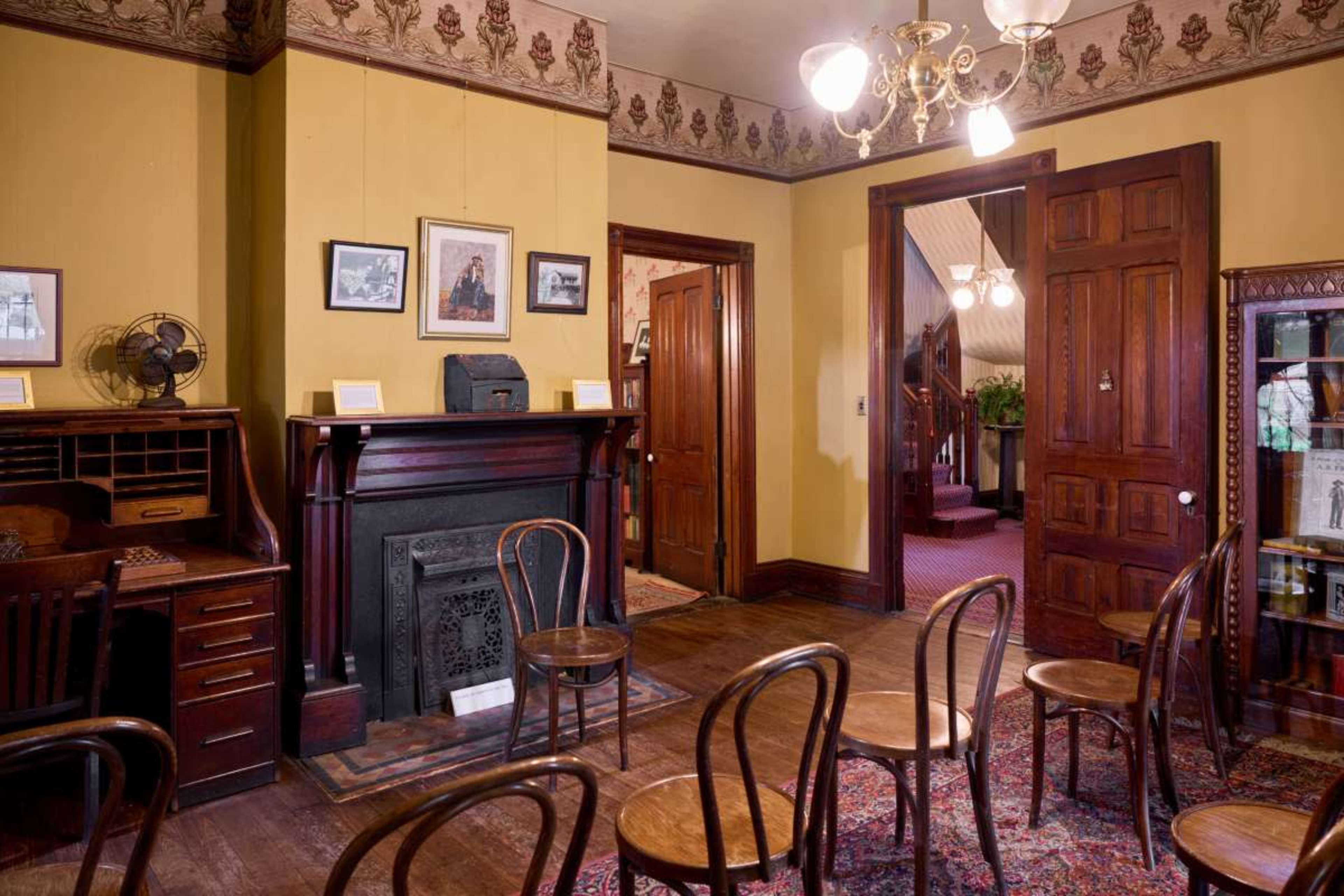 The image shows a furnished room with wooden chairs arranged in rows, a vintage desk and fan against yellow walls, and doorways leading to other areas.