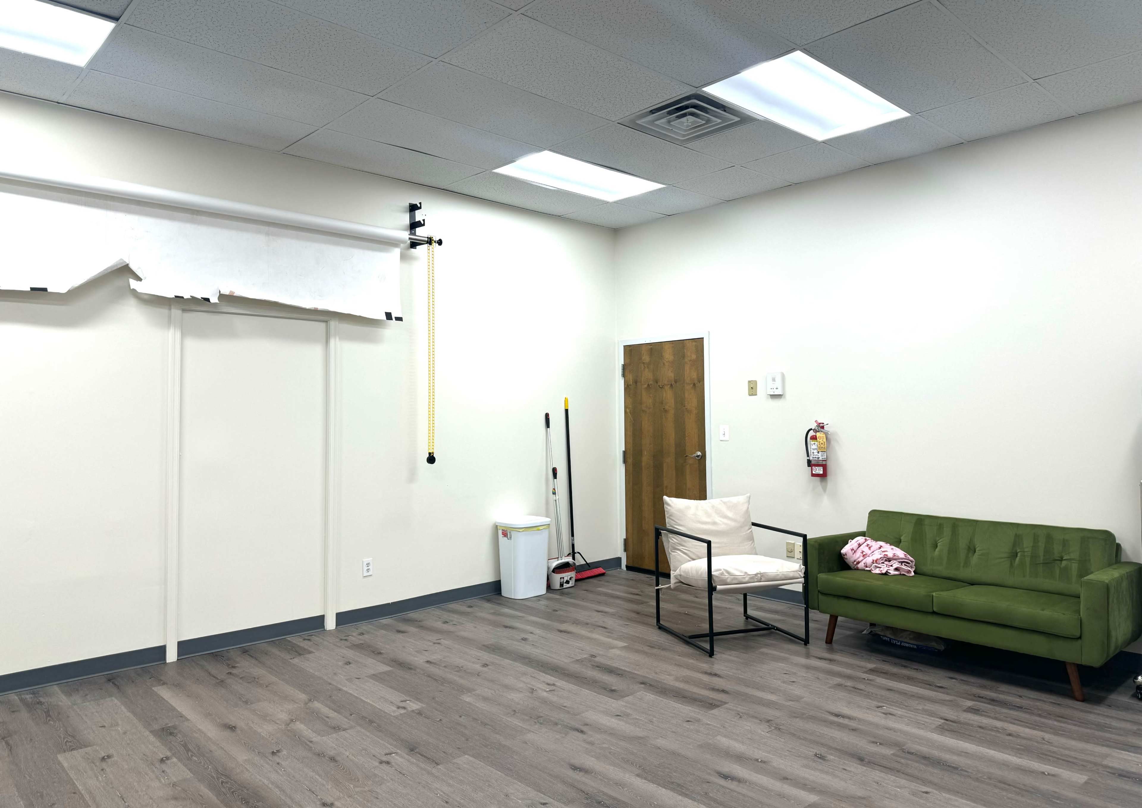A sparse room featuring a green sofa, a black armchair, a wastebasket, and a door on one wall, with a roll of backdrop paper mounted above a plain wall.
