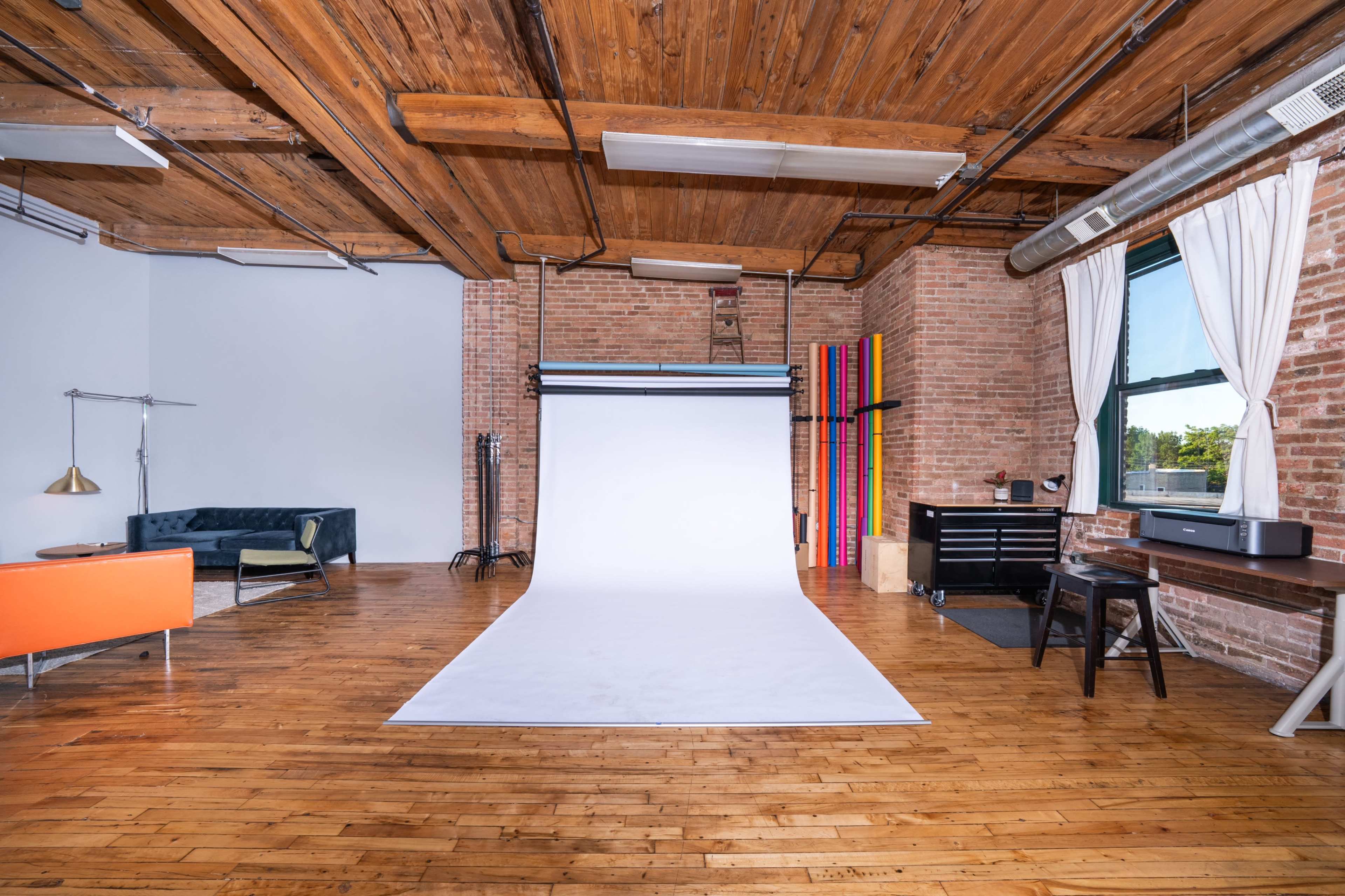 The image shows a spacious photography studio with a white backdrop, wooden floors, exposed brick walls, and various furniture pieces in the corners.