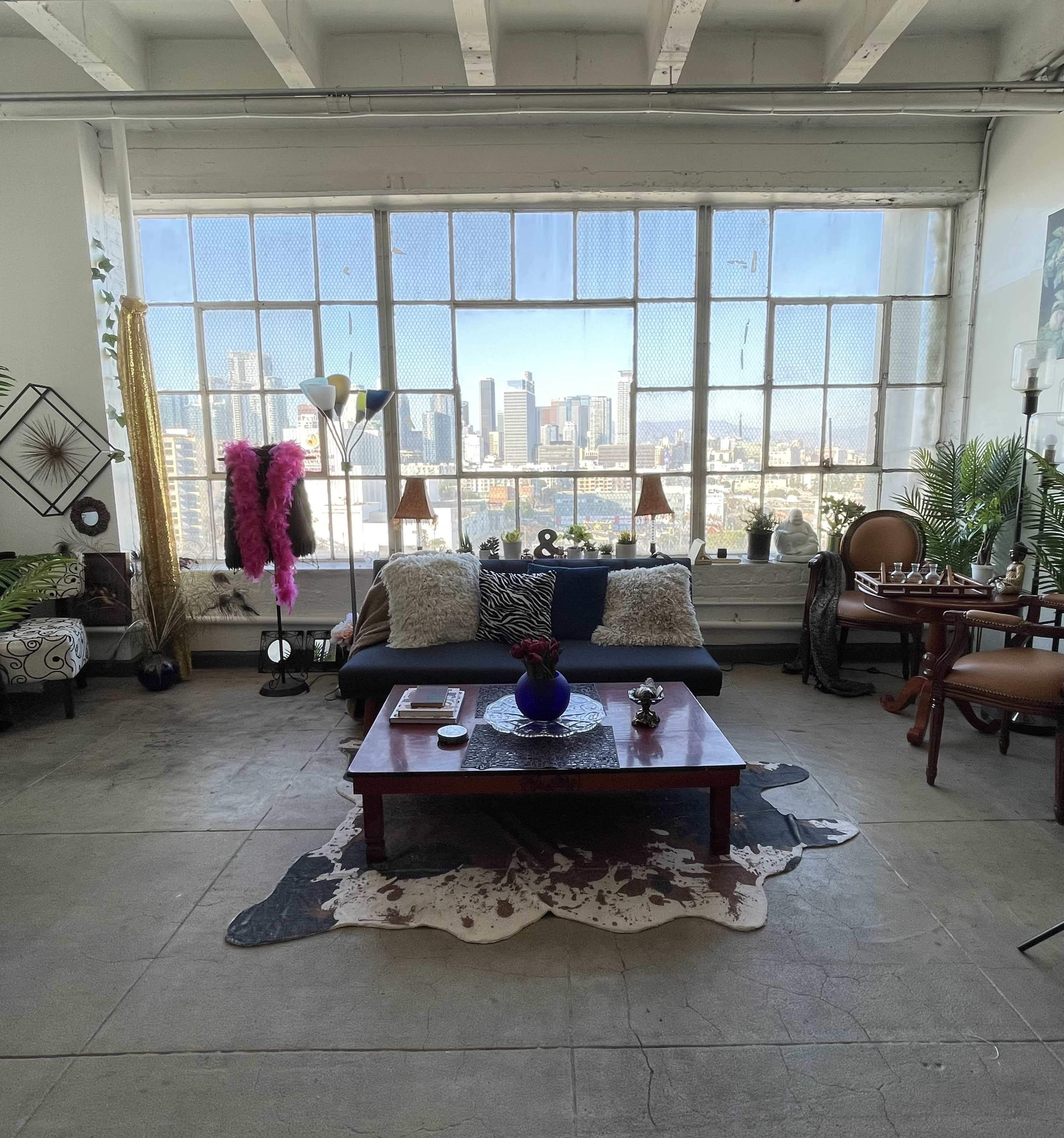 The scene shows a spacious, well-lit living area with a large window displaying a city skyline, featuring a sofa, coffee table, and various decorative plants and furnishings.