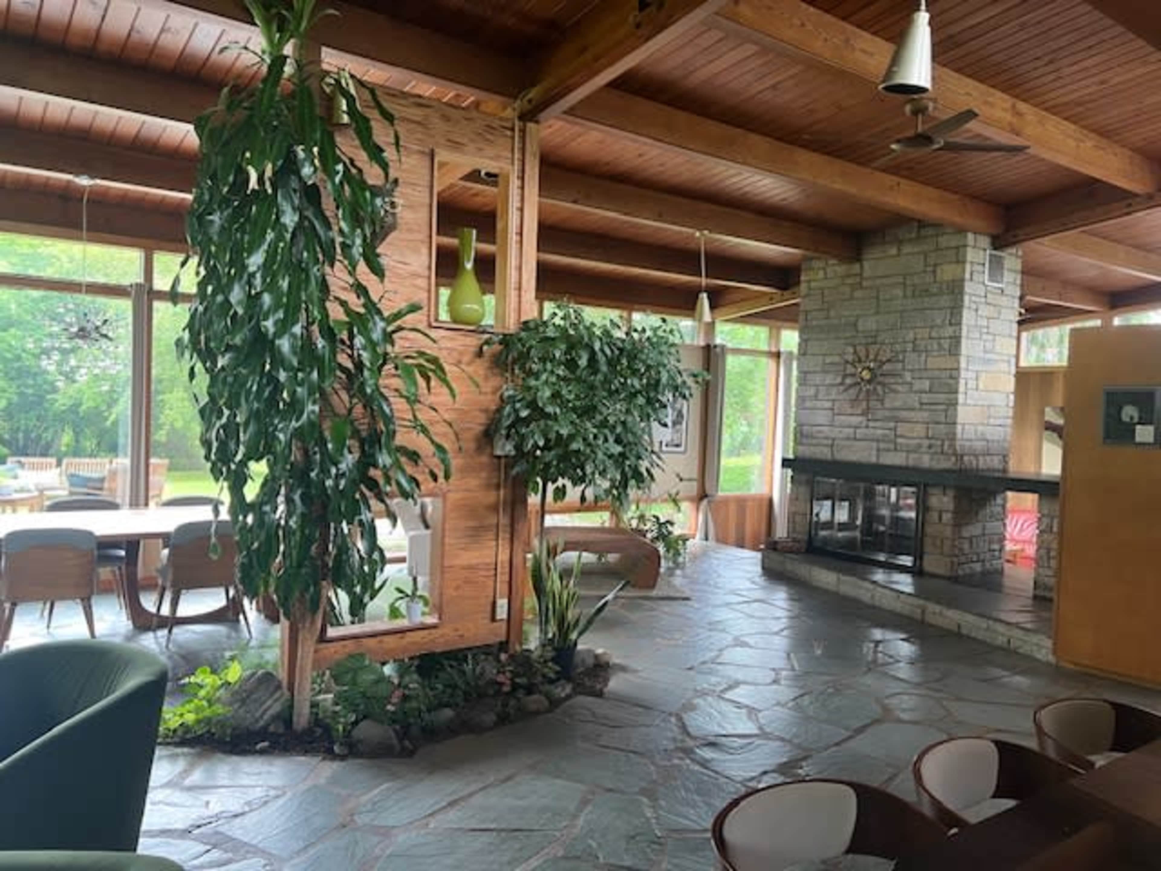 The image shows a spacious interior with wooden beams, stone flooring, and large windows that overlook a green outdoor area.
