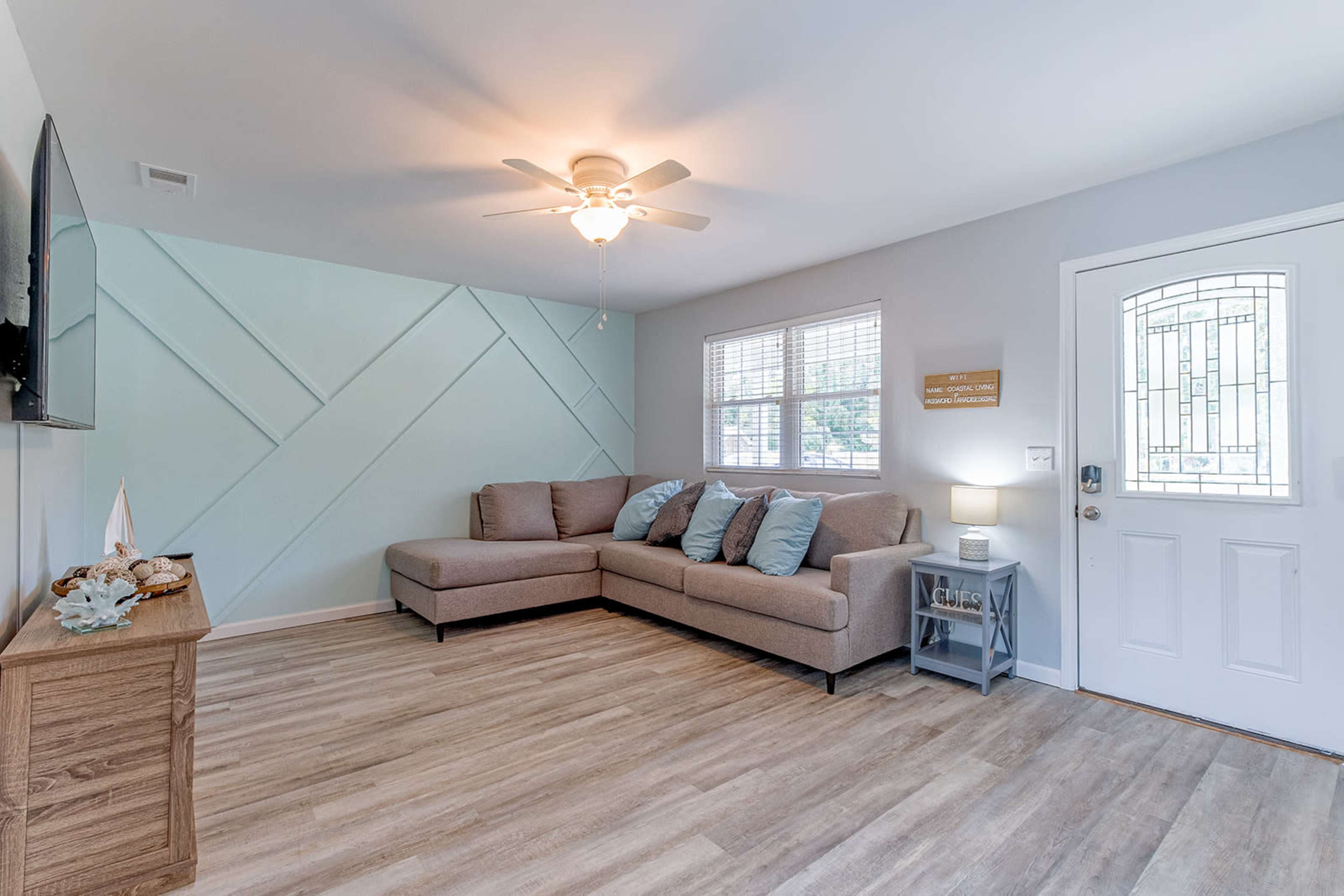 The room features a light-colored sectional sofa with decorative pillows, a wall-mounted television, and a door with decorative glass, all set against a pale blue accent wall.