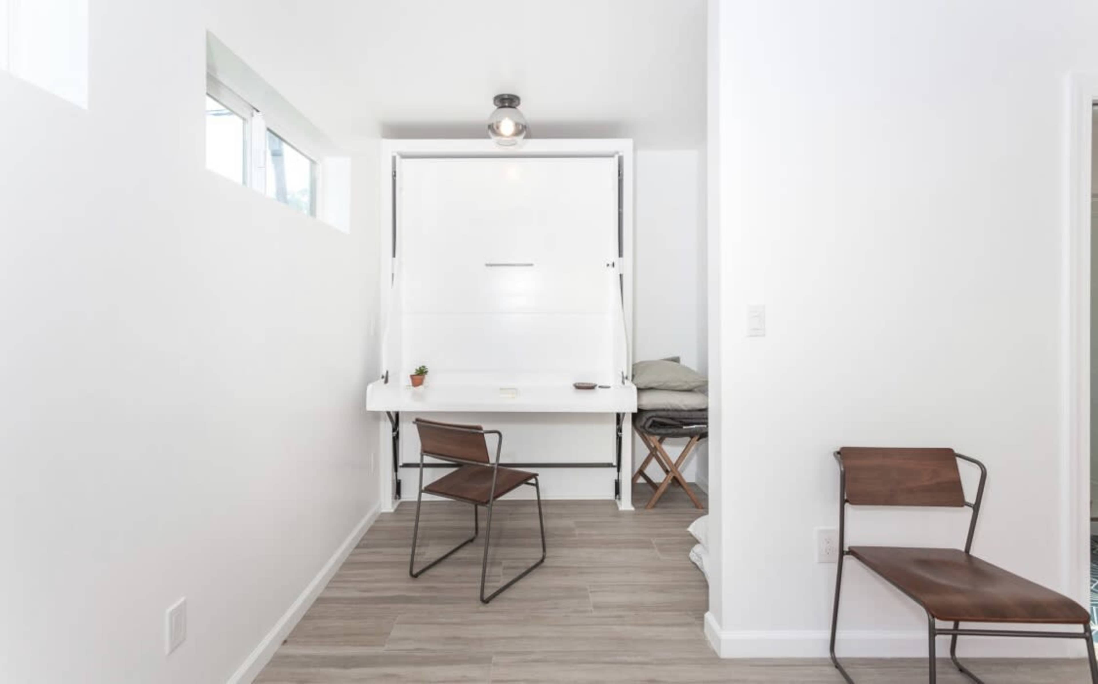 A compact room features a fold-down desk, two metal chairs, and a neatly made bed visible in the background.