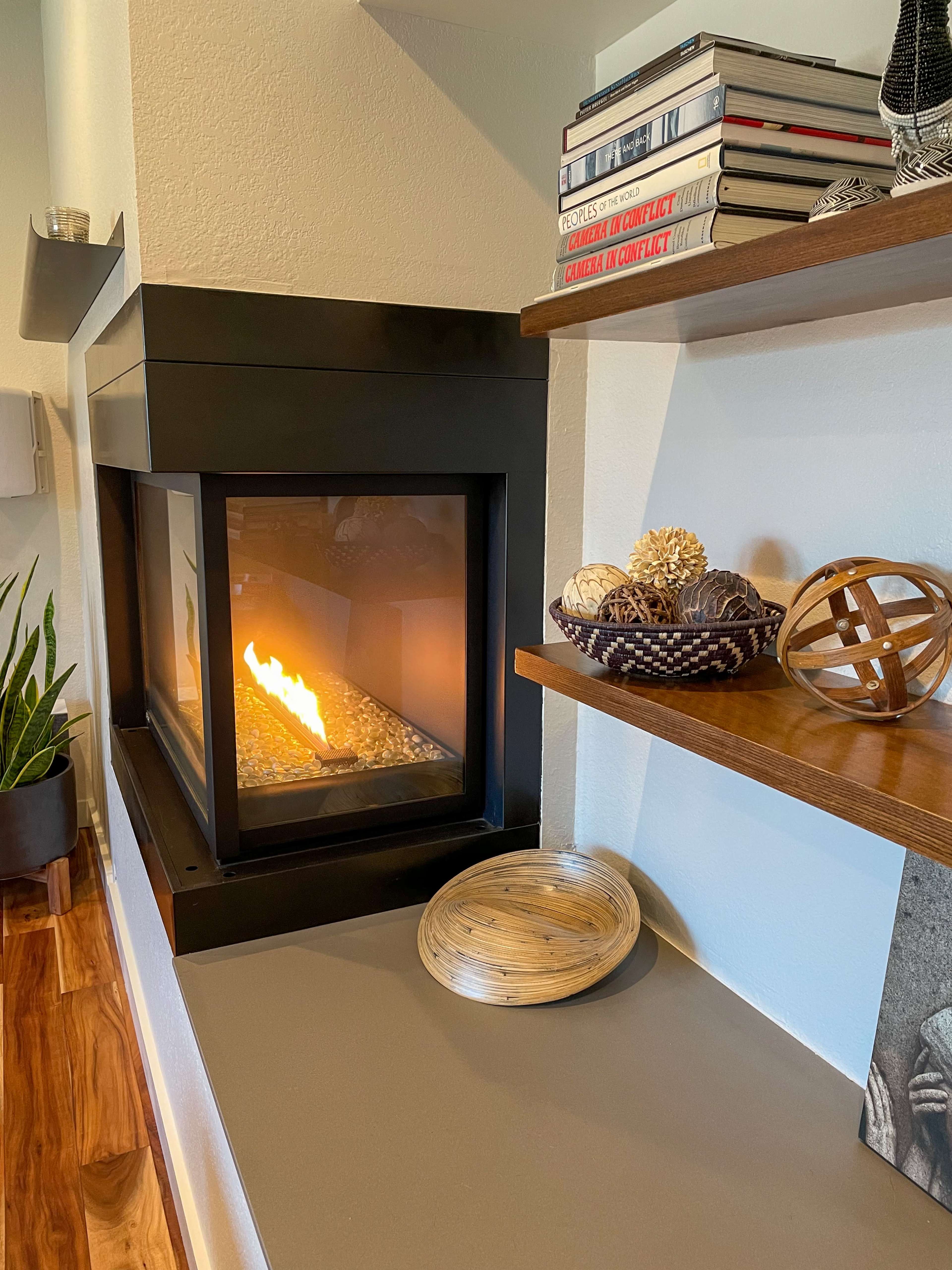 A modern fireplace is set into a wall, with a flame visible through a glass front, accompanied by wooden shelves displaying decorative items and books.