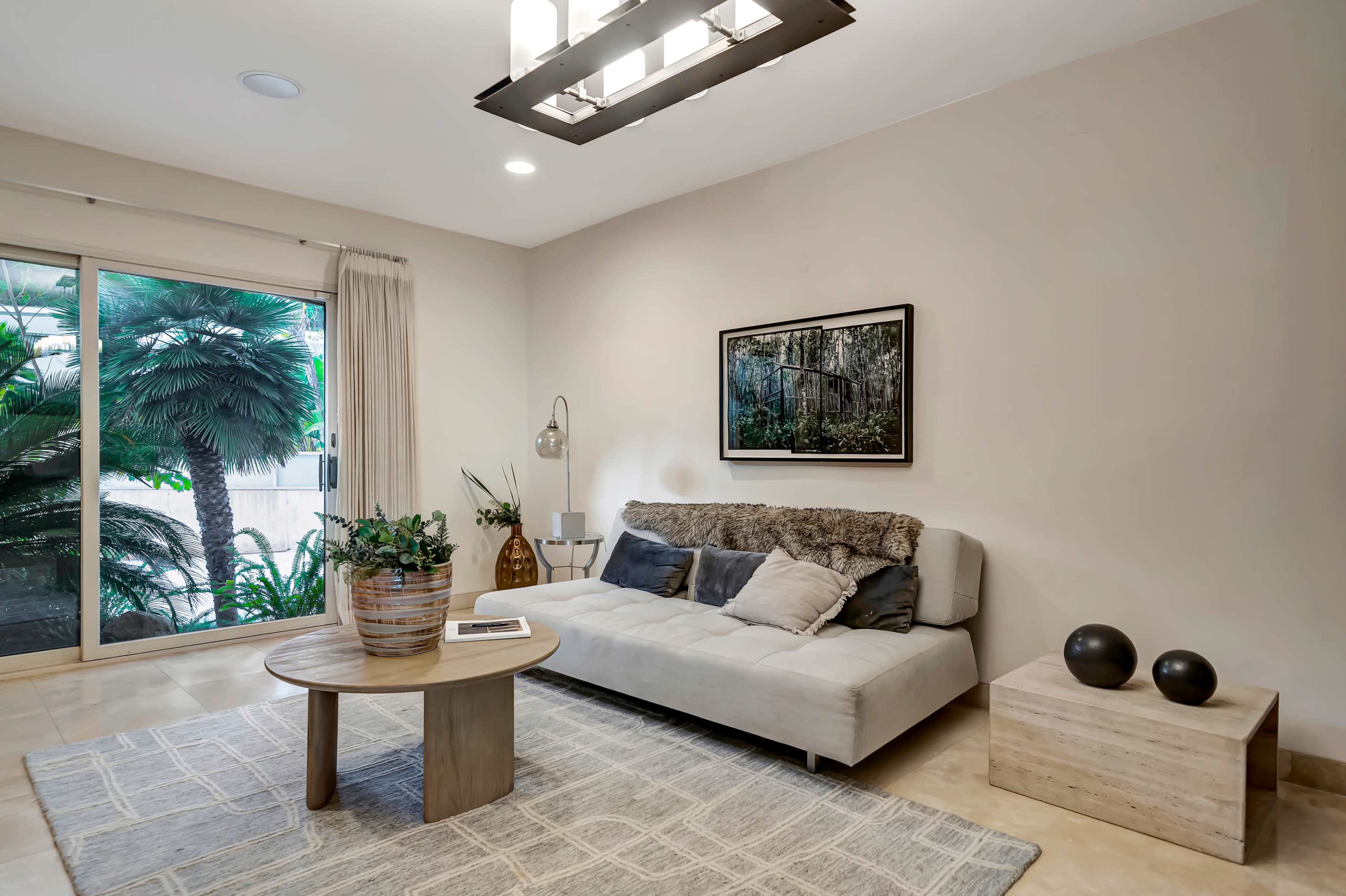 A minimalist living room with a light-colored sofa, a round coffee table, and large windows revealing greenery outside.