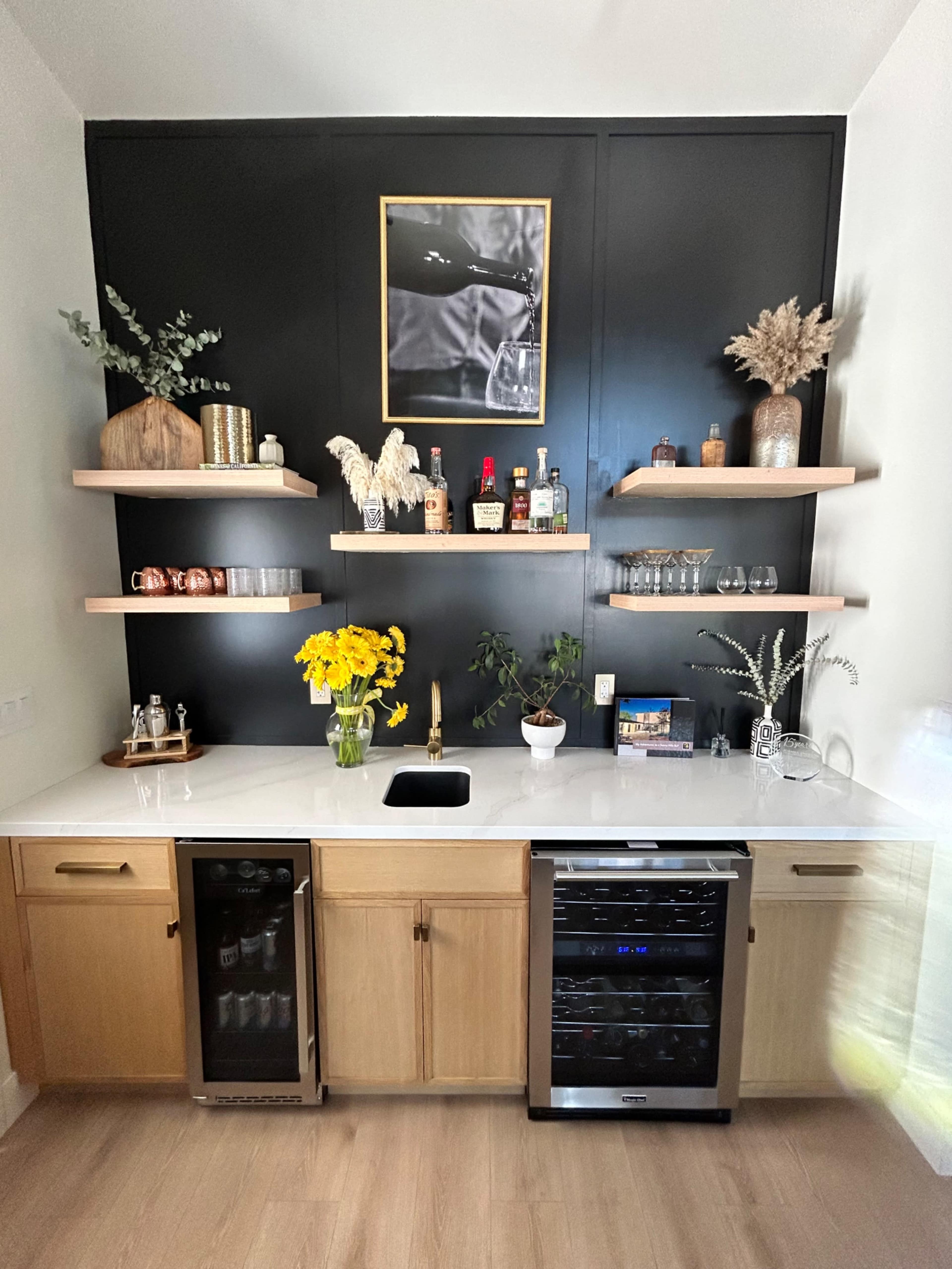 The image shows a modern kitchen bar area with wooden cabinets, a black accent wall, open shelves displaying various bottles and decor, and a sink with a countertop adorned with yellow flowers.