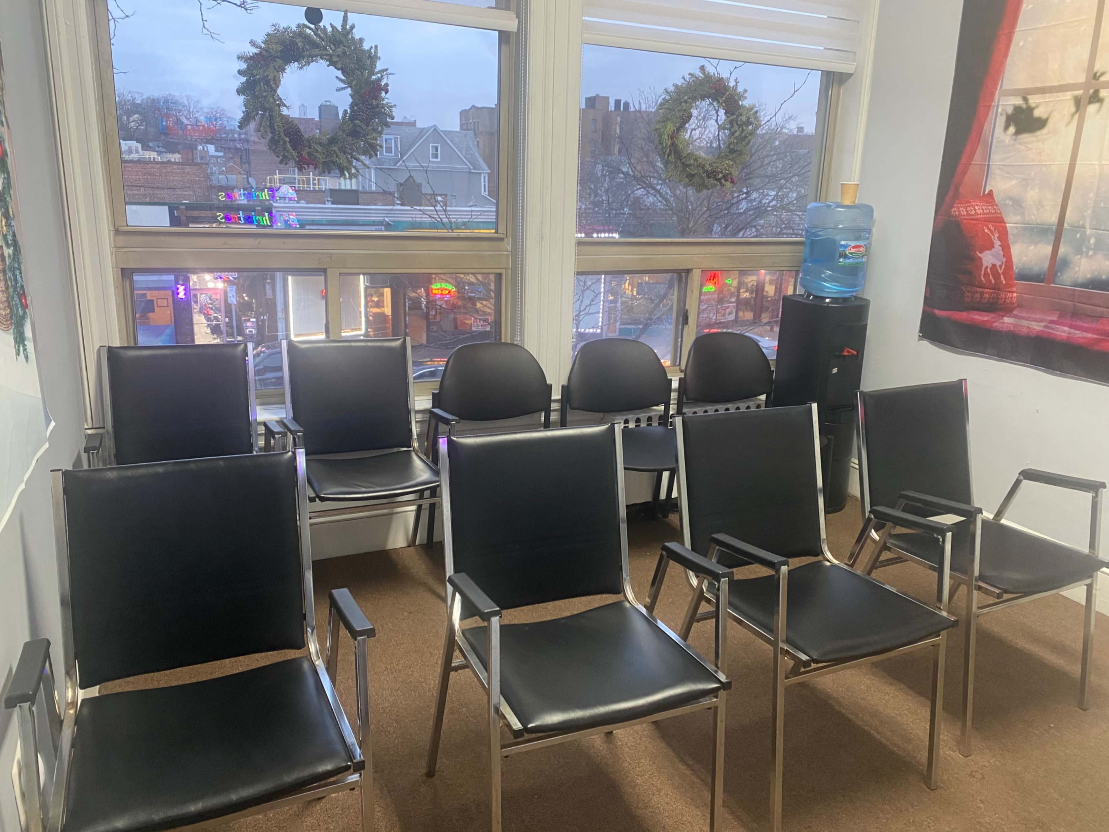 A waiting area with several black chairs arranged in rows facing a window adorned with wreaths, overlooking a city street.