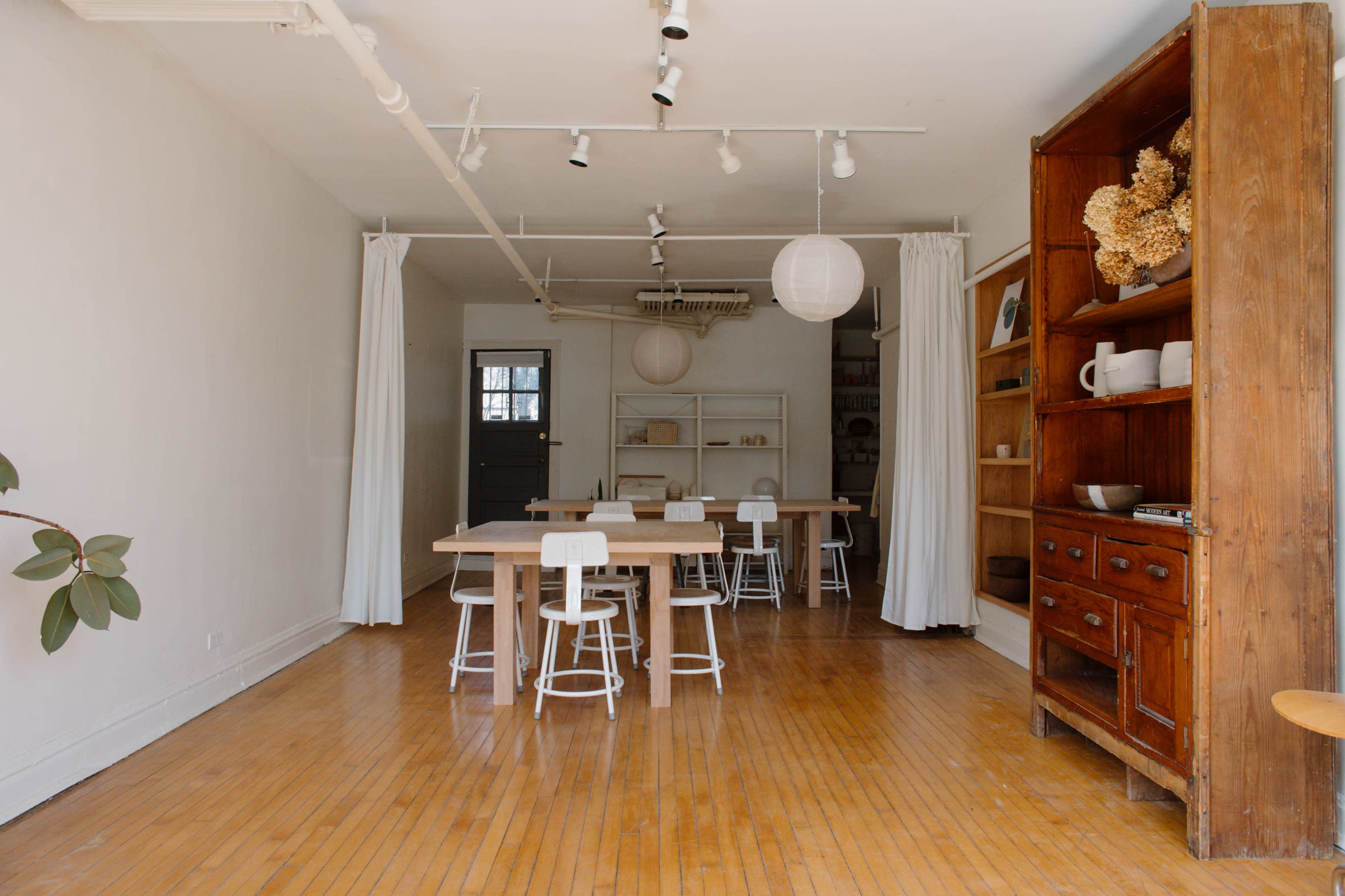 The image shows a spacious, well-lit room with wooden floors, featuring several tables and chairs, along with a wooden storage unit against one wall.