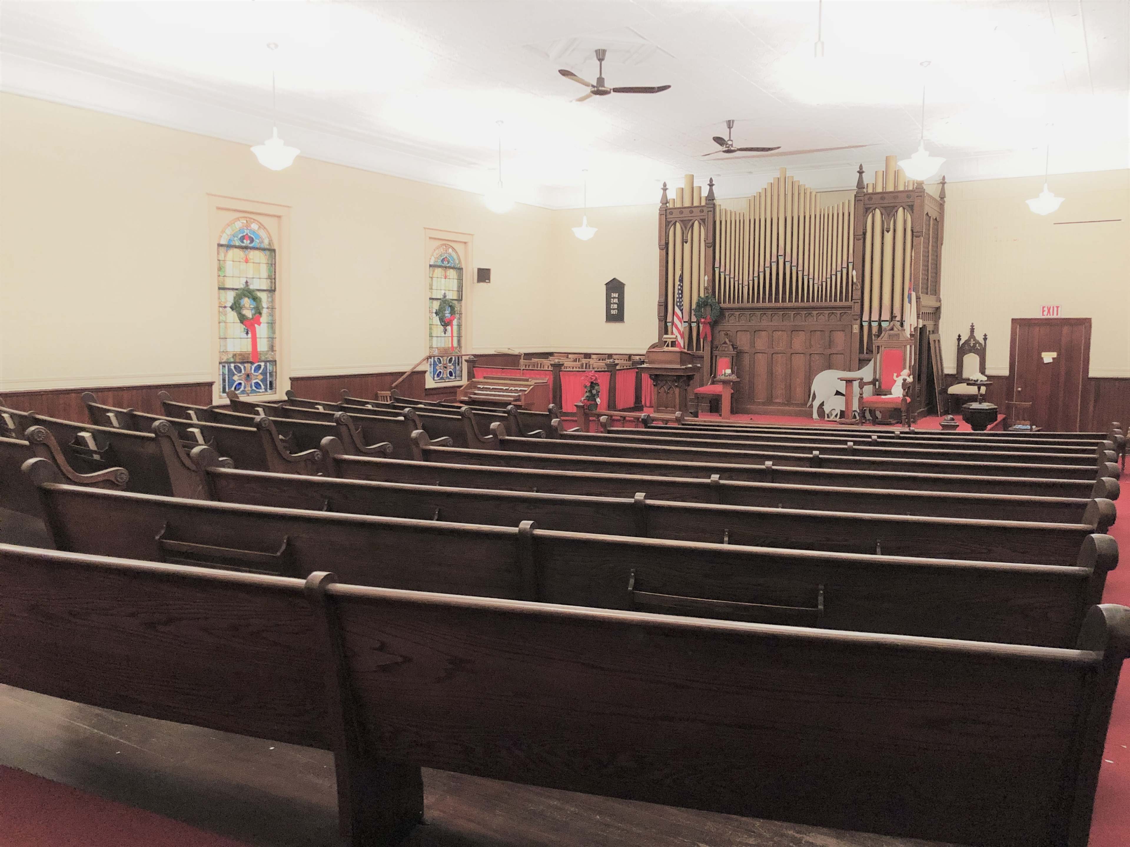 A church interior with wooden pews facing a podium and a large pipe organ in the background, flanked by stained glass windows.