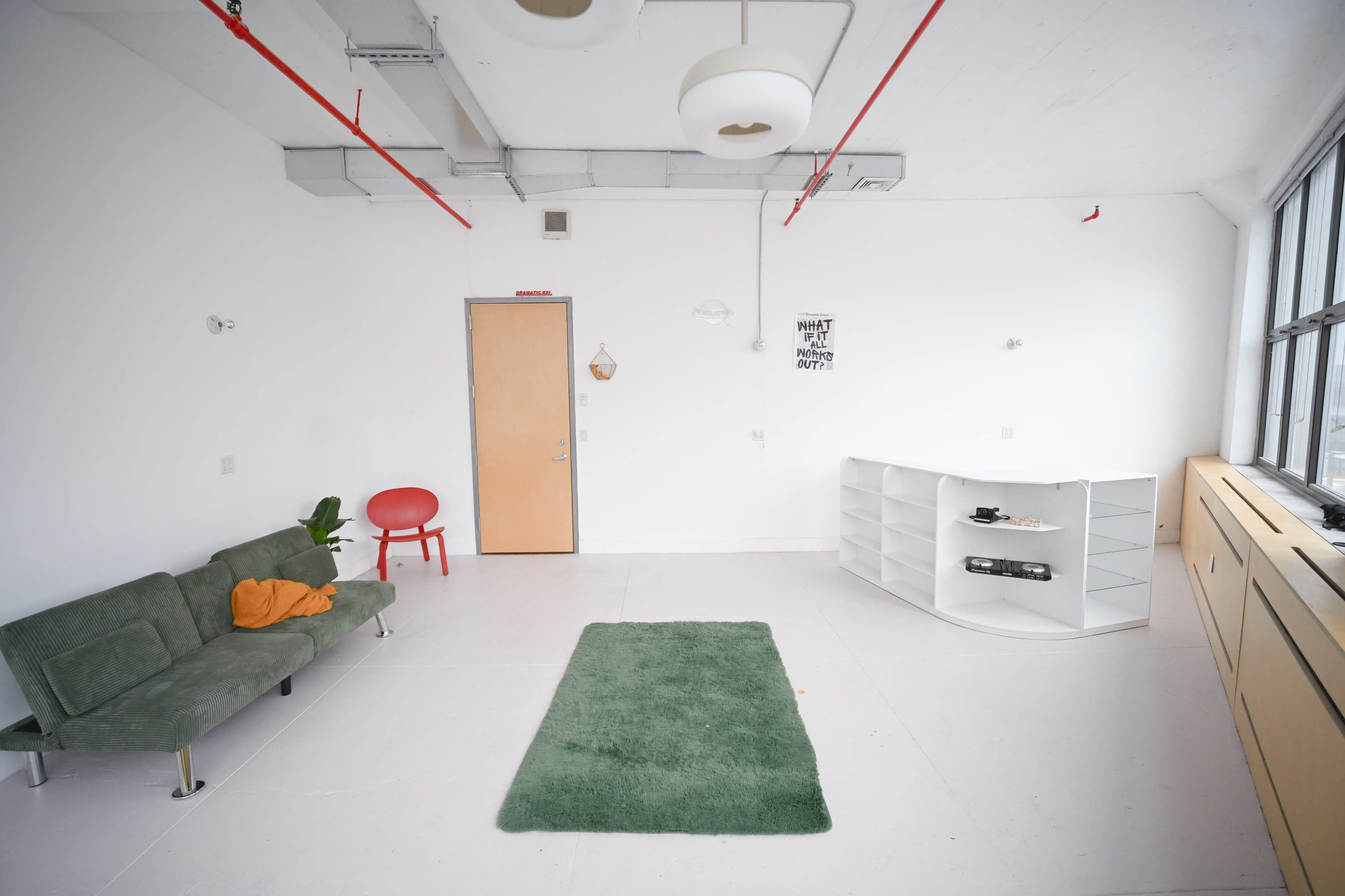 The room features a green rug in the center, a gray sofa with an orange throw, a red chair, a white shelving unit, and a door.