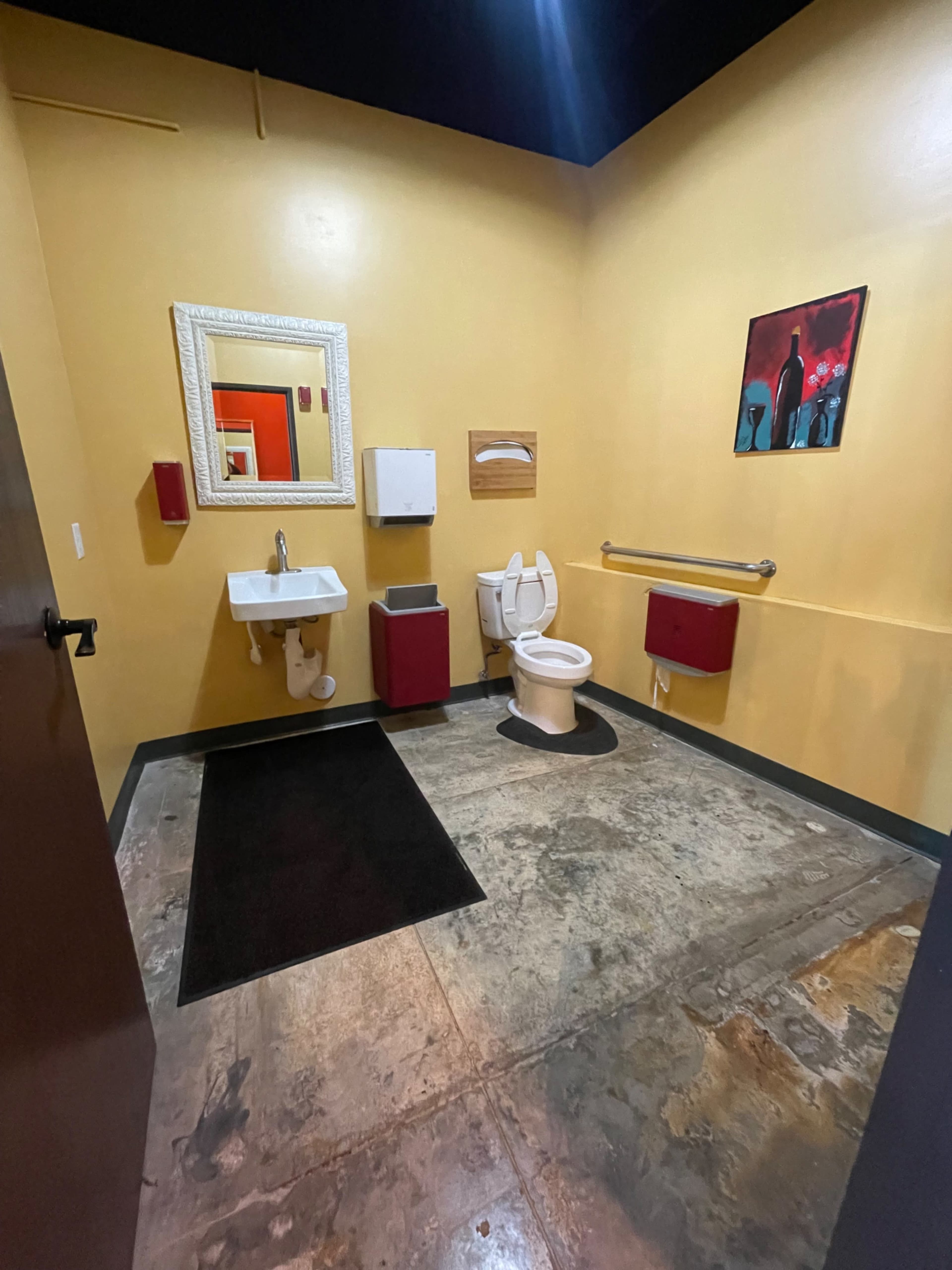 A restroom with a toilet, sink, wall-mounted hand dryer, and a large mirror, all set against a yellow wall and featuring a concrete floor.