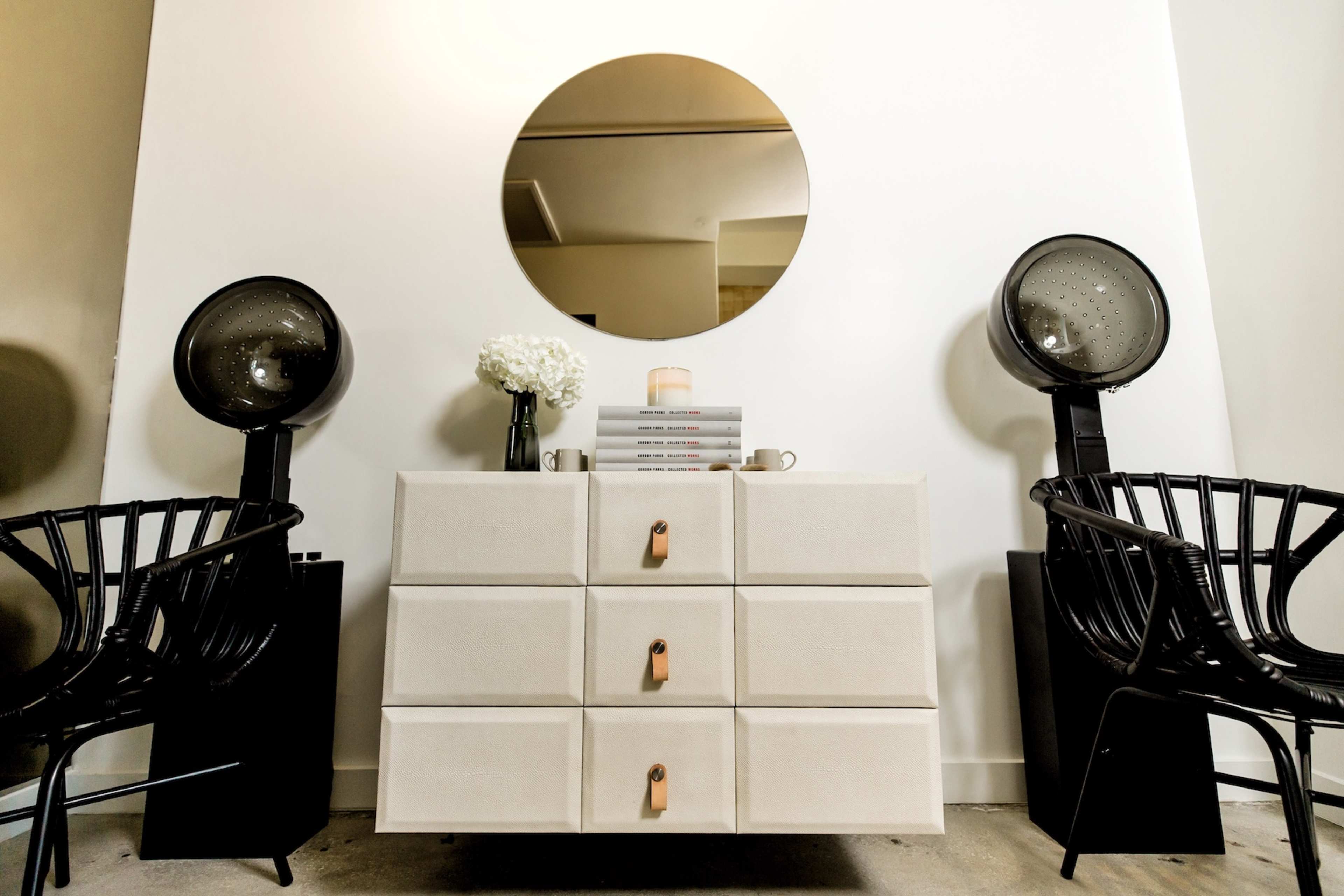 A light-colored dresser with four drawers, flanked by two black chairs and a round mirror above it, with a vase of flowers and a stack of books on the dresser.