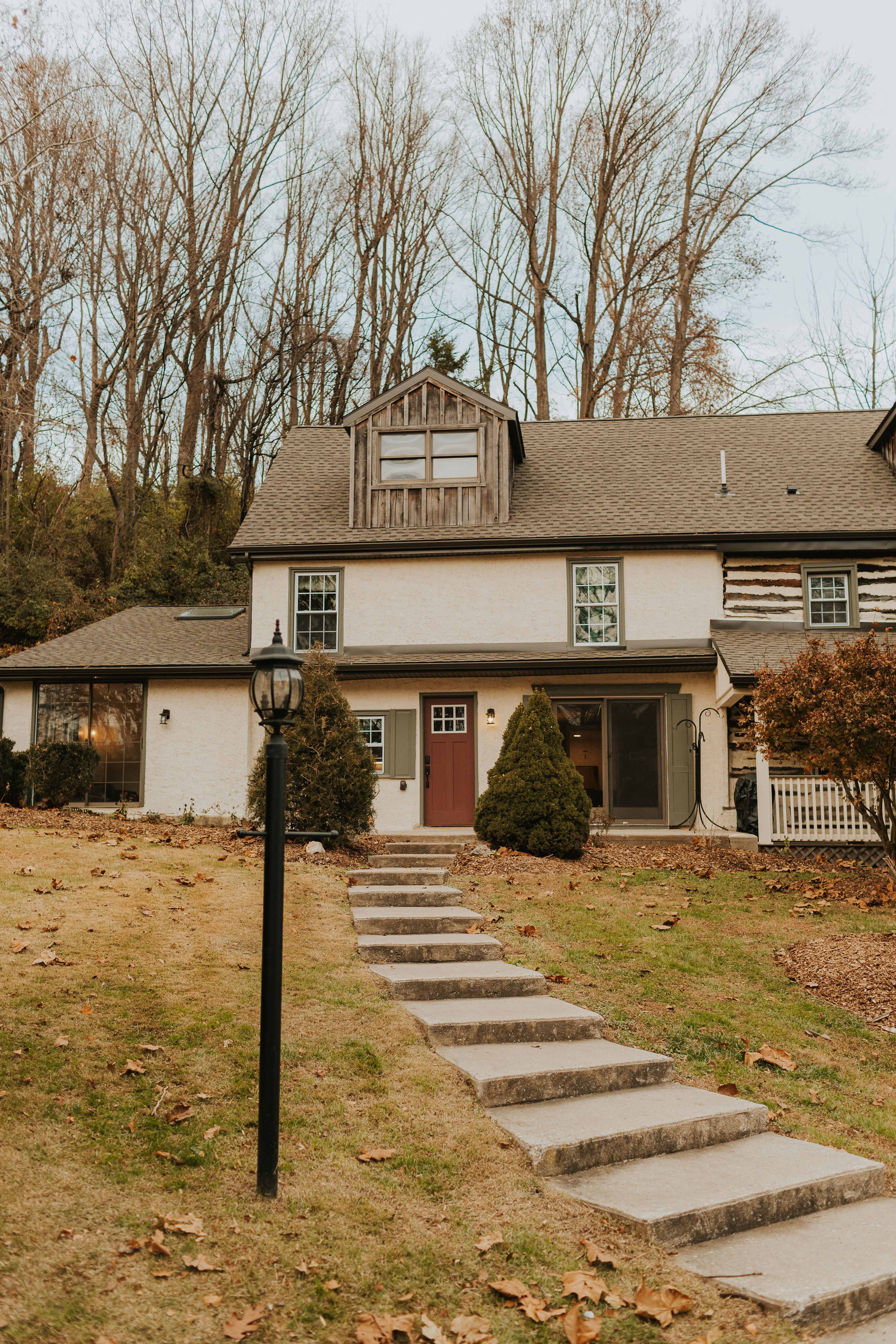 A two-story house with a combination of log and painted walls is situated at the end of a stone pathway lined with grass and trees.