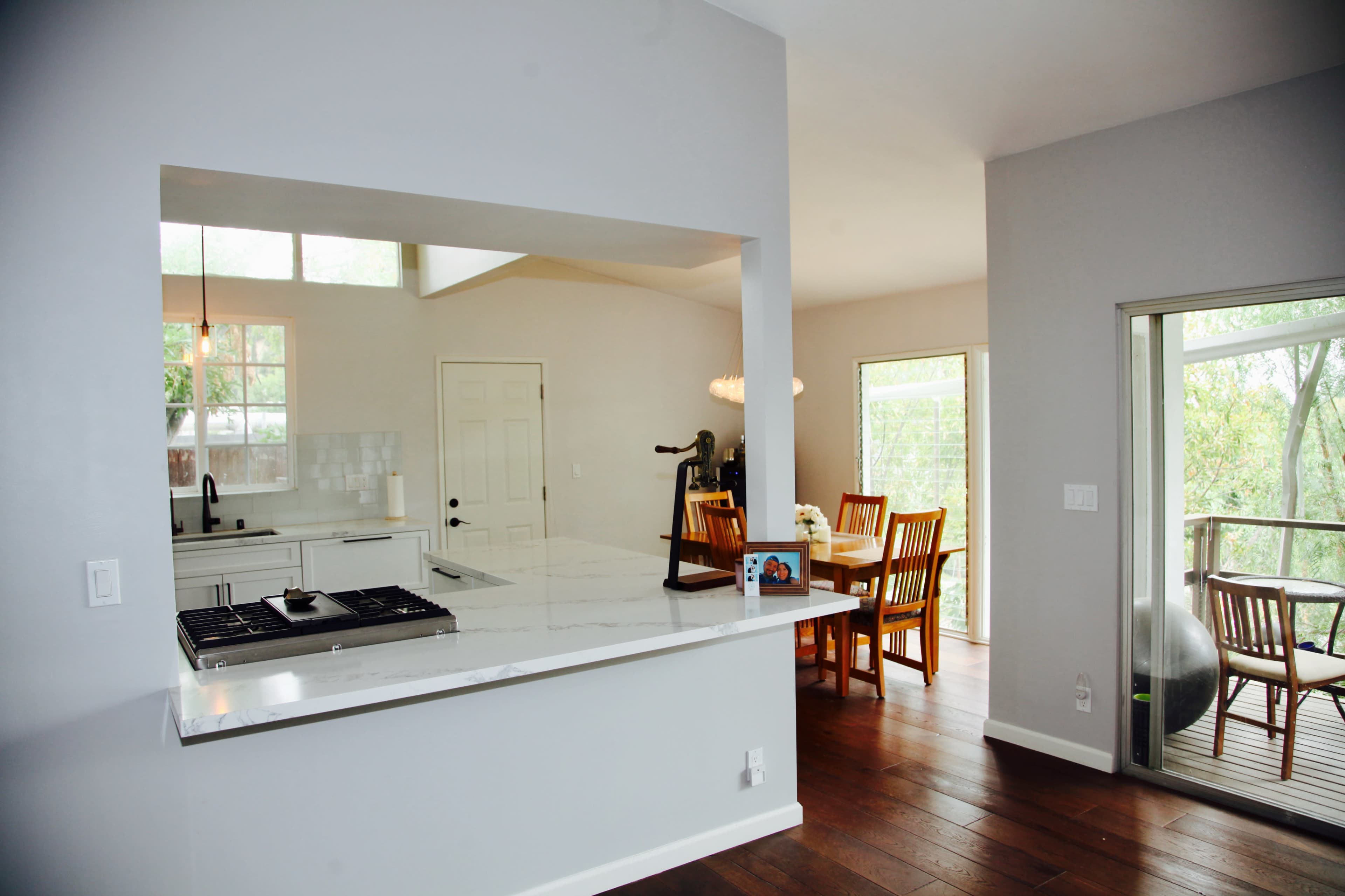 The image shows a modern kitchen with a white countertop and a view of a dining area and a deck through large windows.