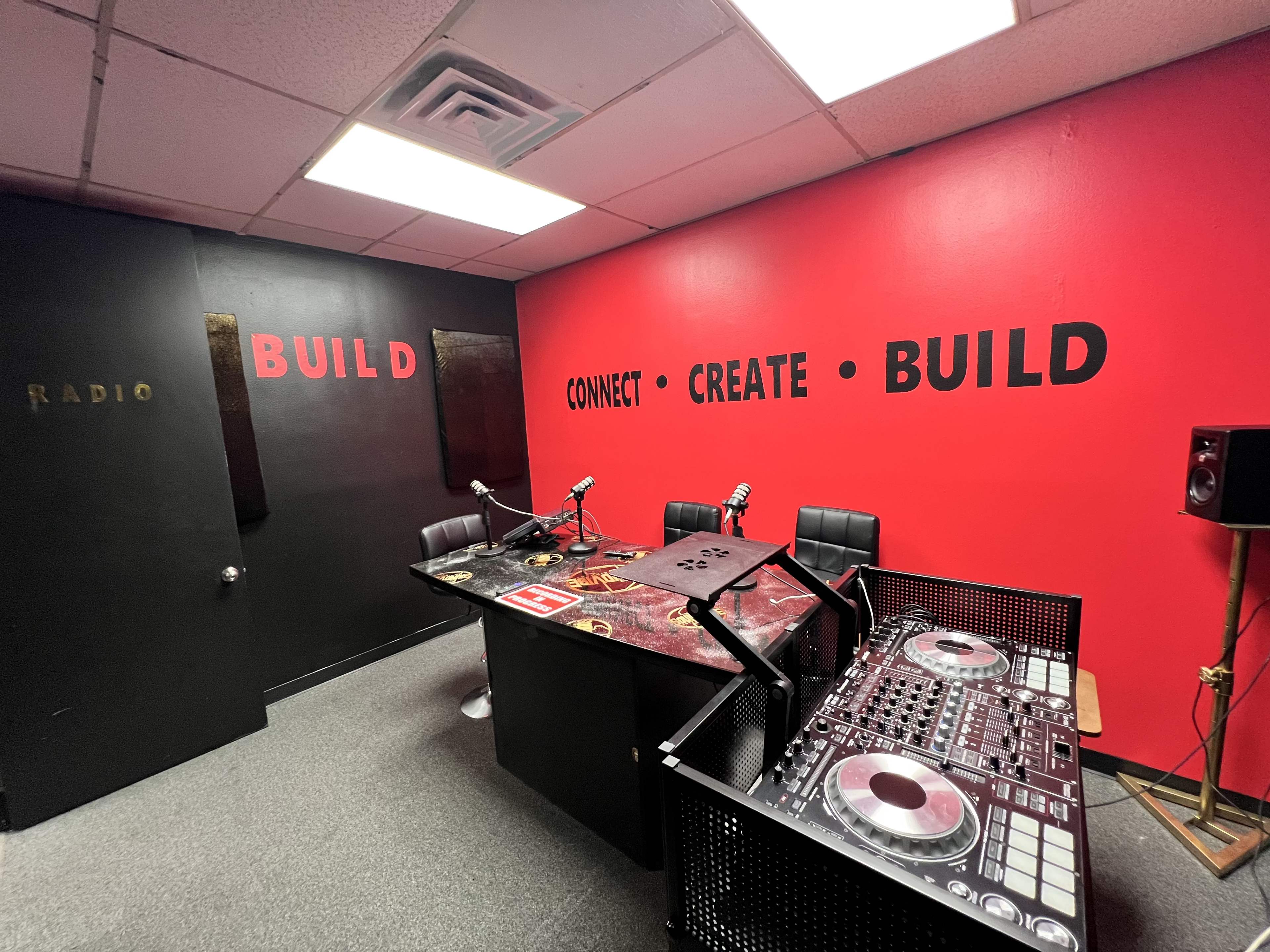 A studio with a red accent wall featuring the words "CONNECT," "CREATE," and "BUILD," alongside a black table equipped with microphones and a DJ setup.