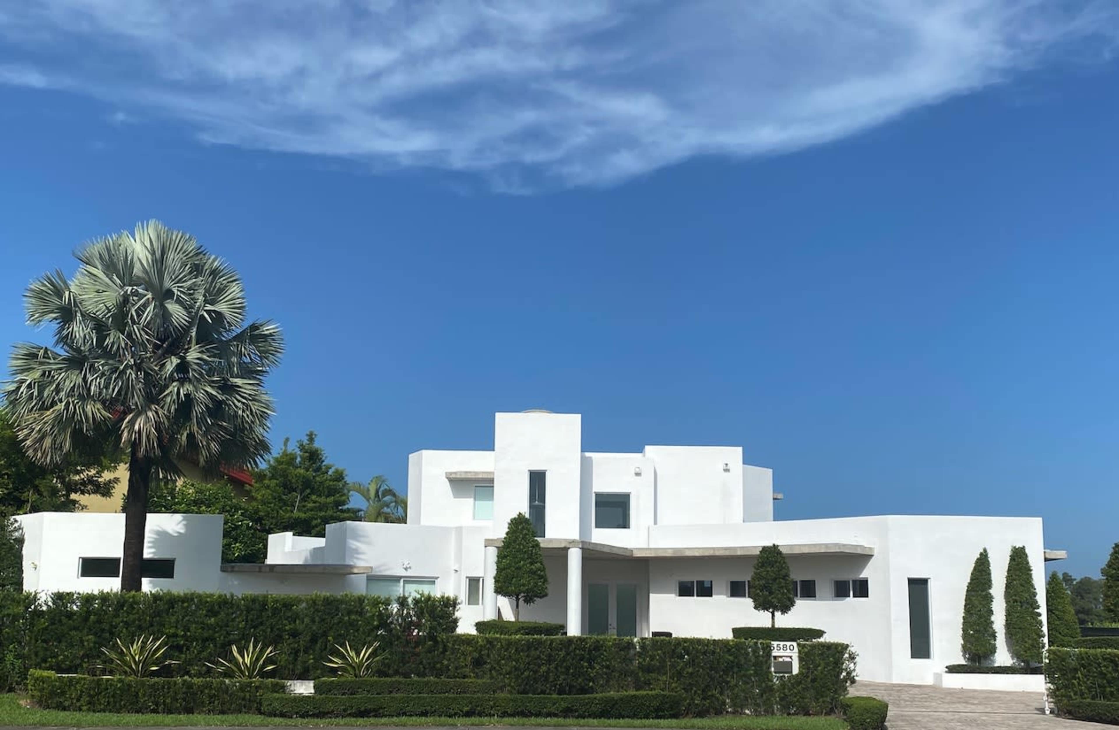 A modern white house with geometric architecture is surrounded by neatly trimmed hedges and palm trees under a clear blue sky.