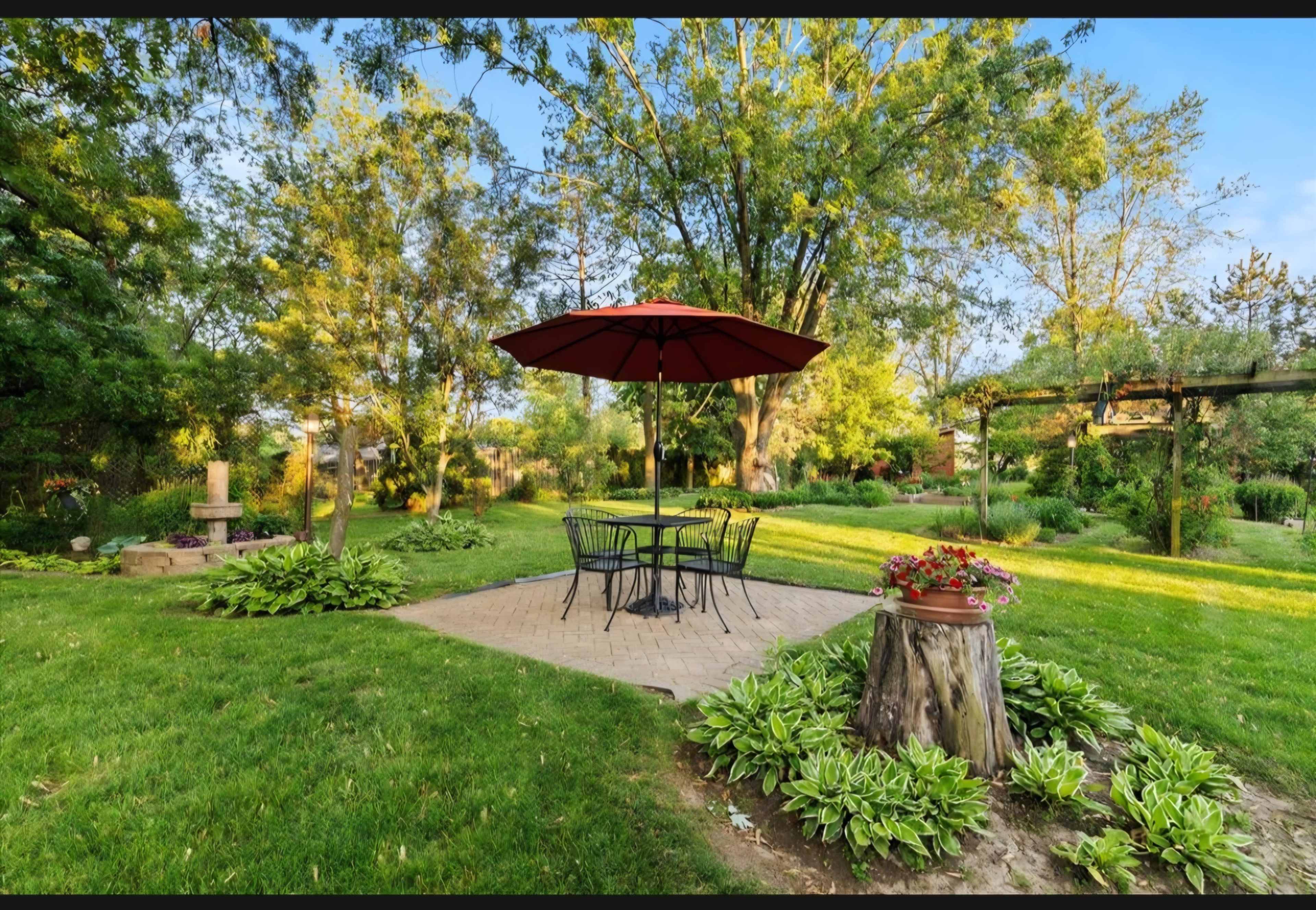 A circular patio with a red umbrella and black chairs sits surrounded by lush greenery and trees.