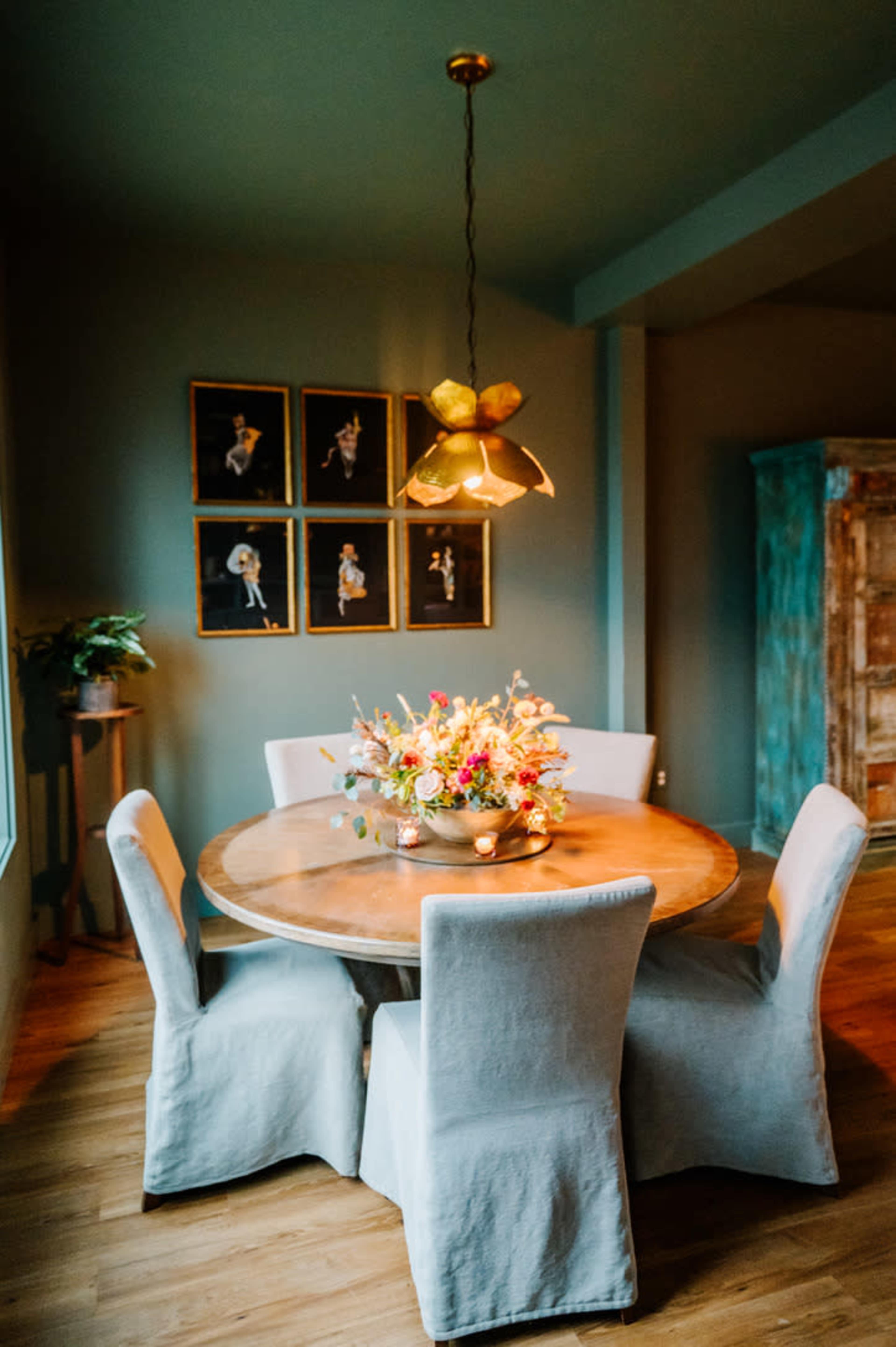 A round wooden dining table with four upholstered chairs is set beneath a pendant light, surrounded by floral decor and framed artwork on the walls.