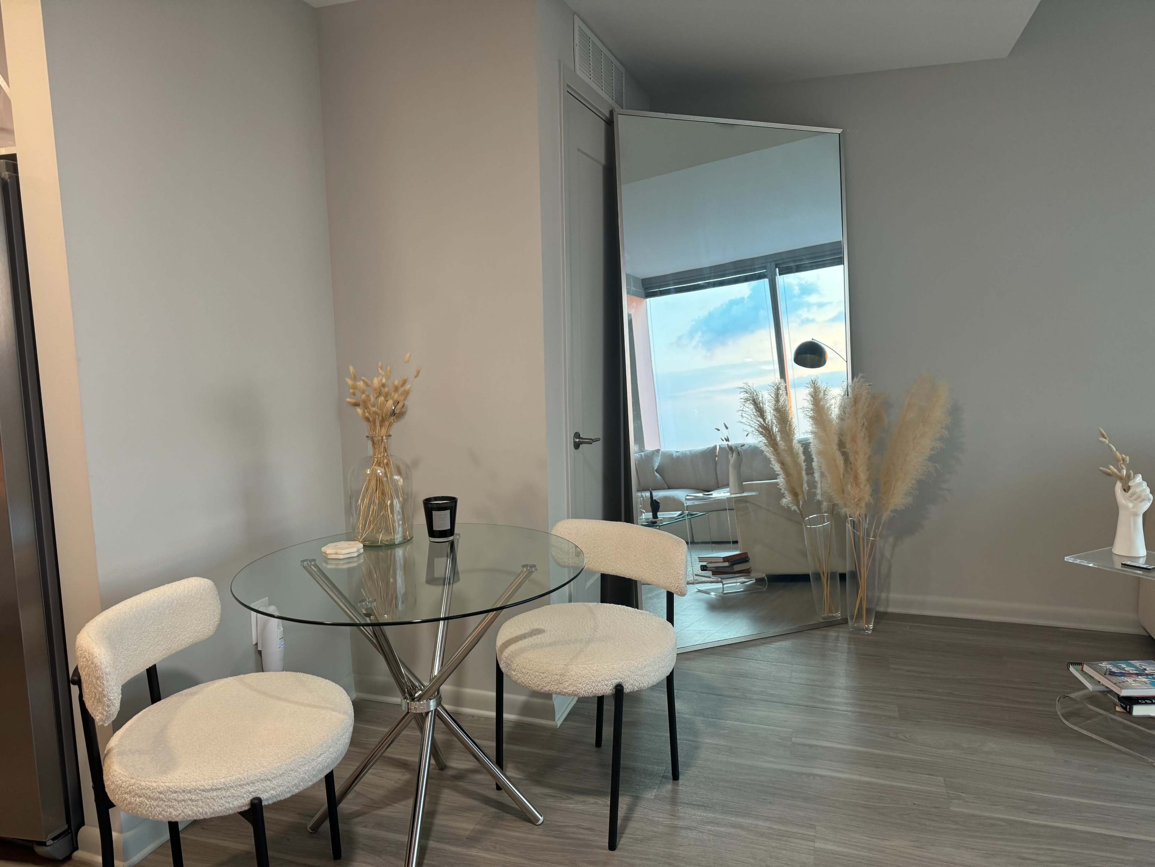A minimalist dining area features a glass table with two upholstered chairs and a large mirror reflecting the space.