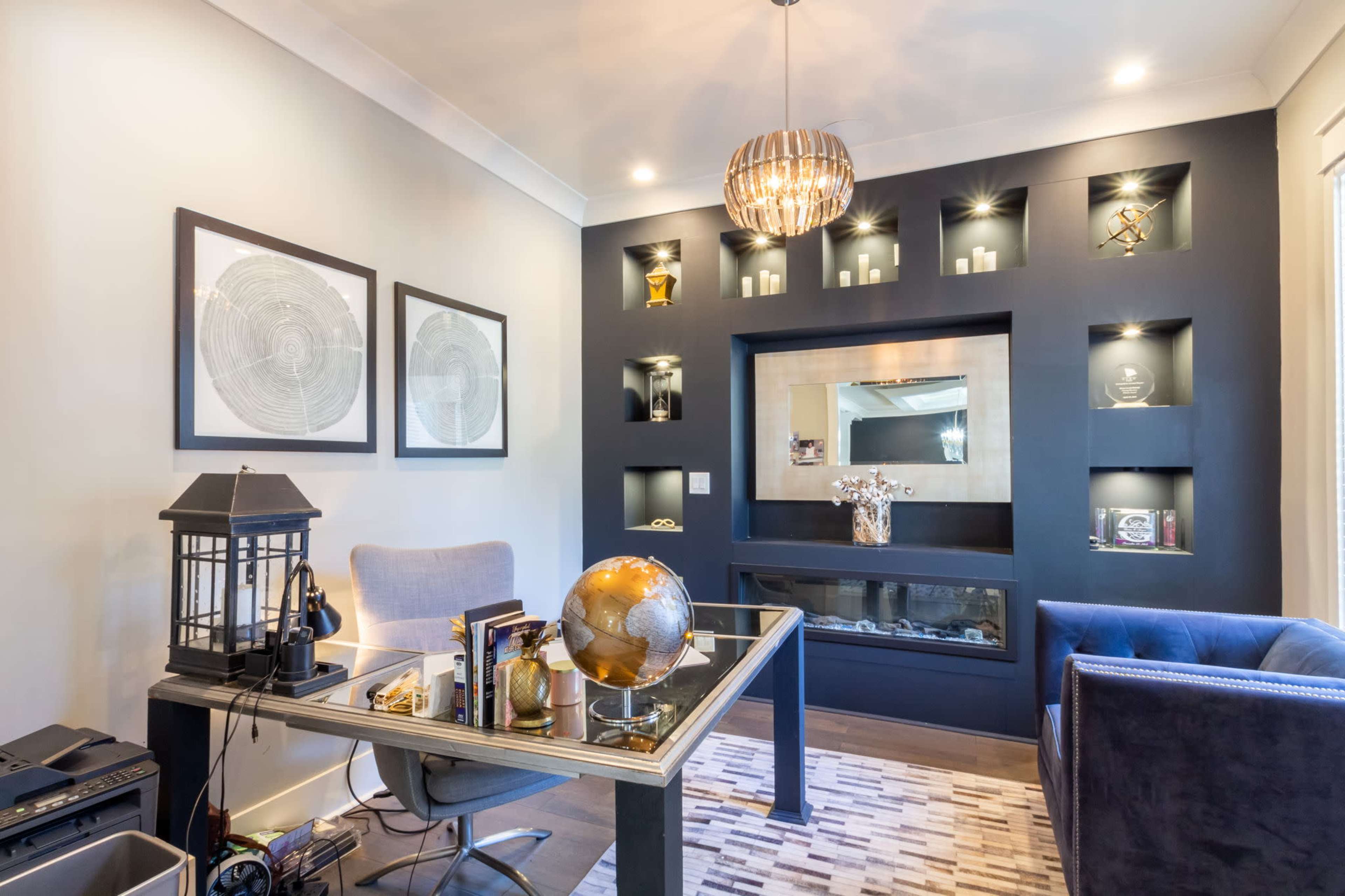 A modern office features a glass-top desk with a globe and decorative items, accented by dark walls with shelves showcasing various objects and framed artwork.