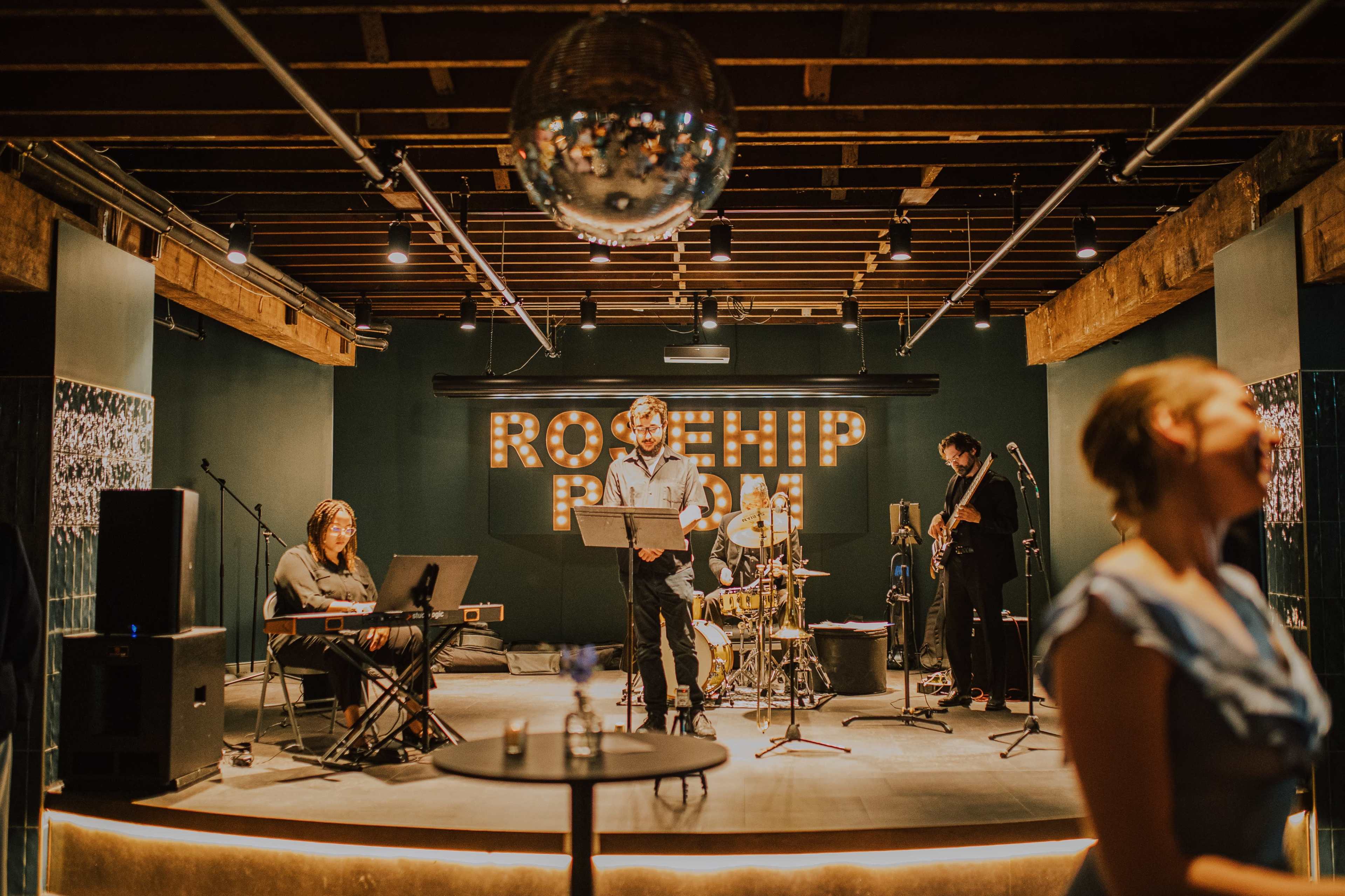 A live band performs on stage in the Rosehip Room, with a keyboardist, vocalist, and drummer amid a modern venue setting.