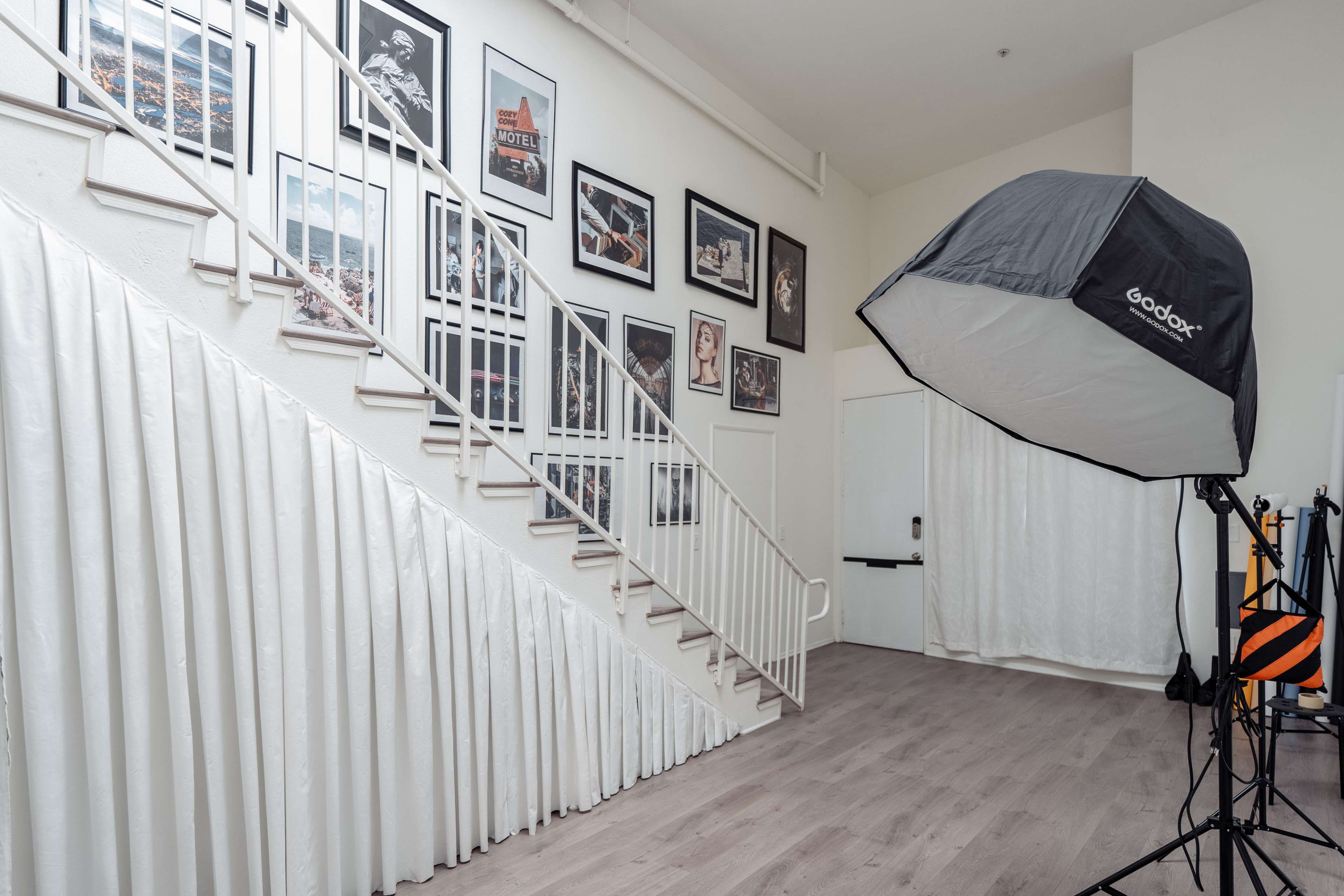 A staircase with a white railing is flanked by framed photographs on the wall, and a photography light is positioned near a white curtain door.