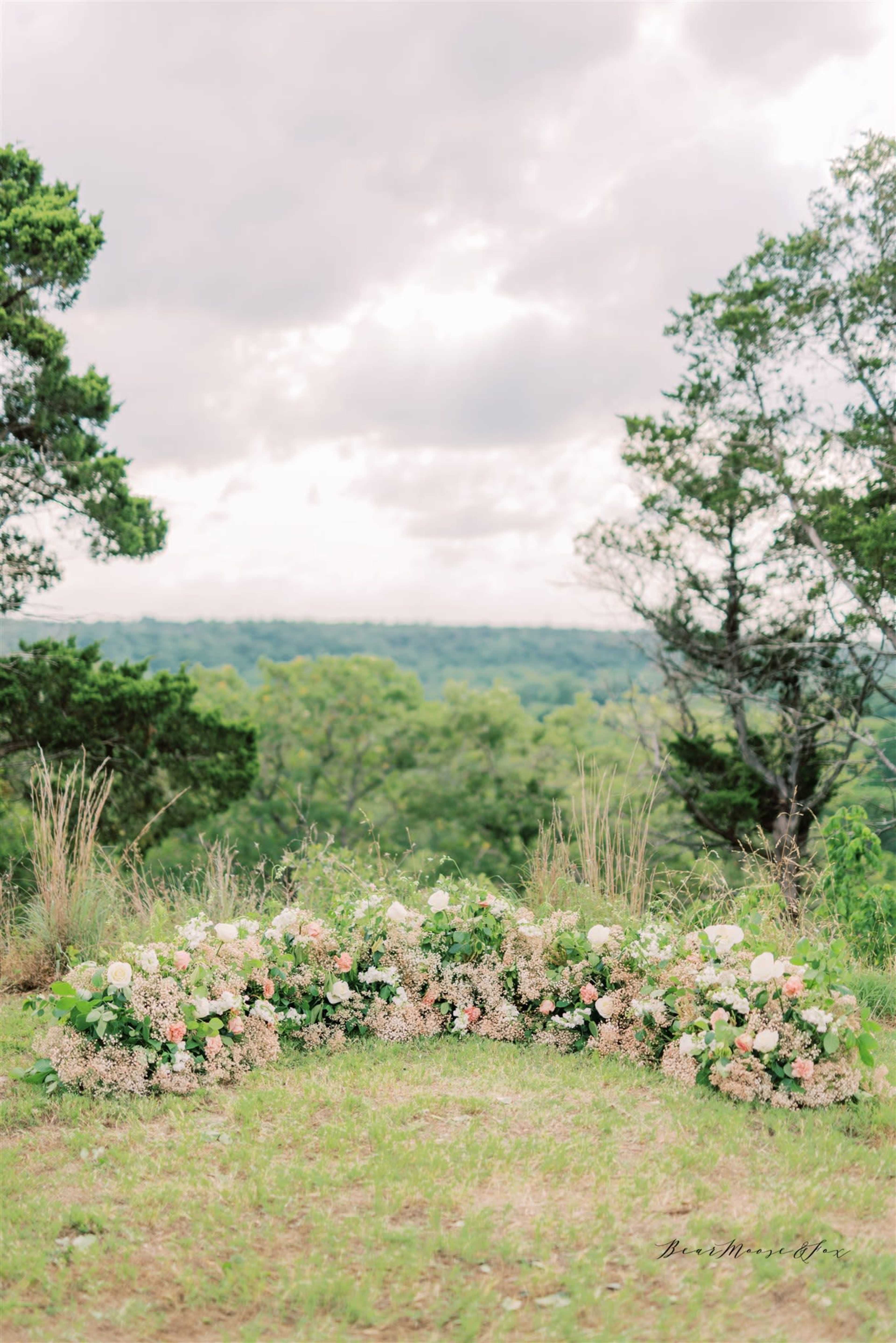 A floral arrangement with pink and white blooms is displayed in a grassy area overlooking a green landscape and cloudy sky.