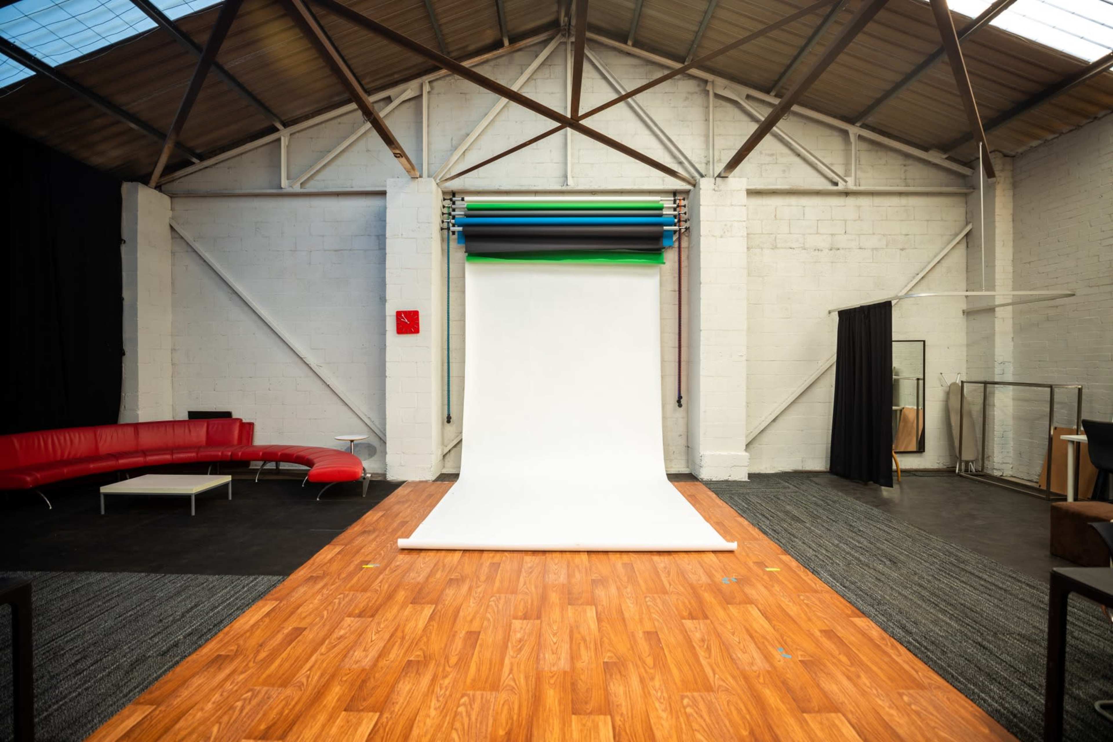 A spacious photography studio with a white backdrop, wooden flooring, a red couch, and black curtains.