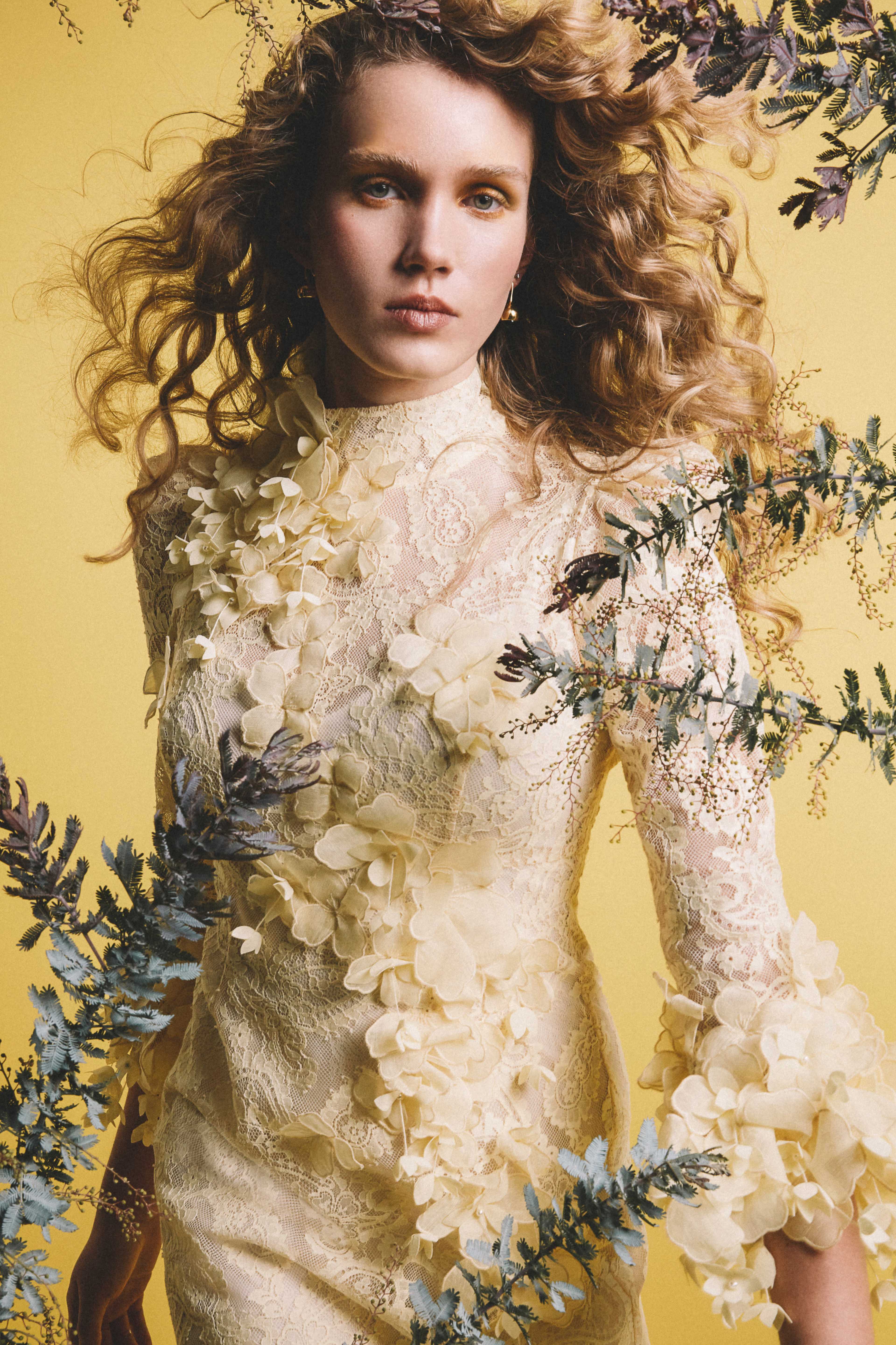 A model wearing a cream-colored lace dress stands against a yellow background, surrounded by floral arrangements.