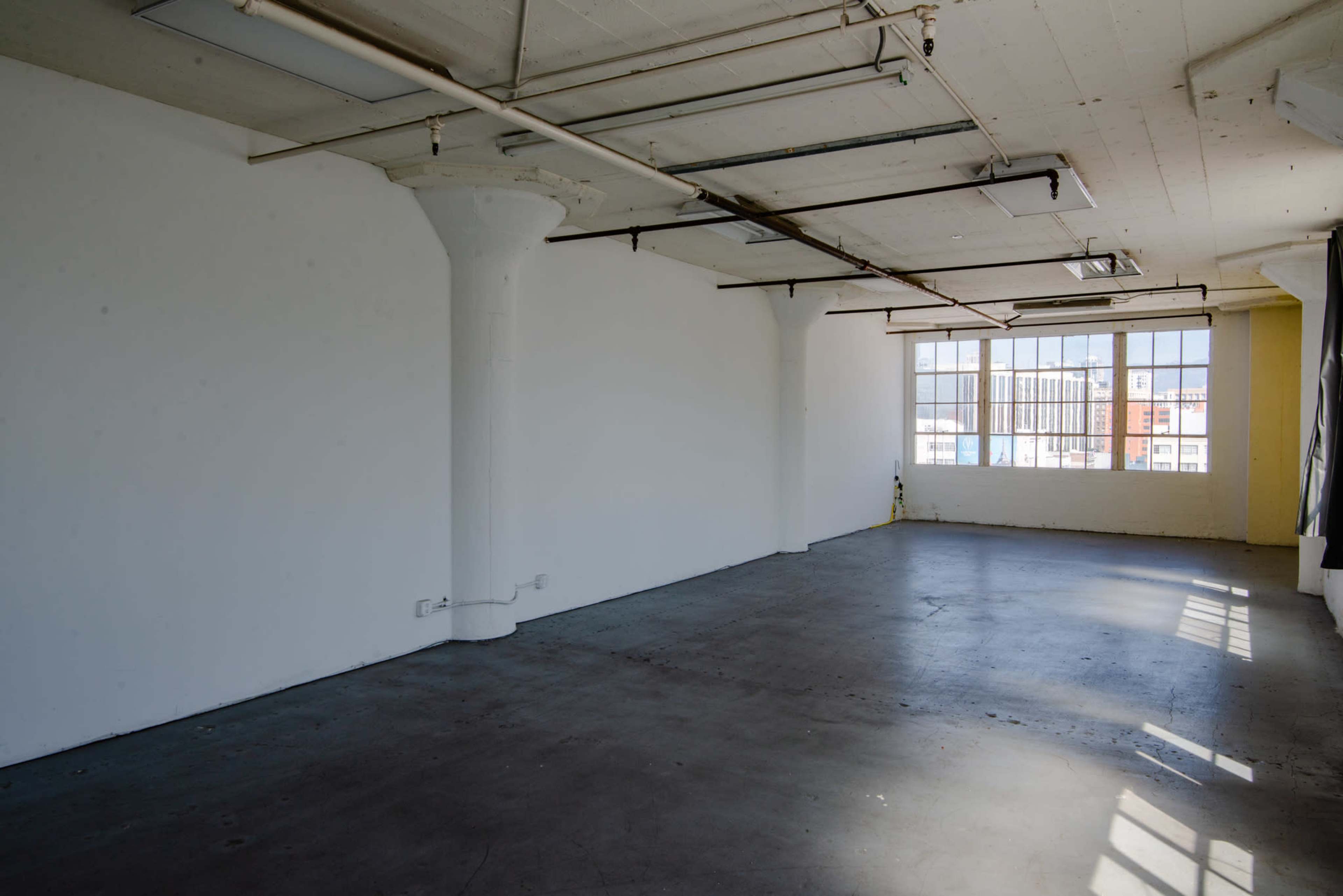 The image showcases an empty industrial-style room with concrete floors, large windows, and exposed ceiling beams.