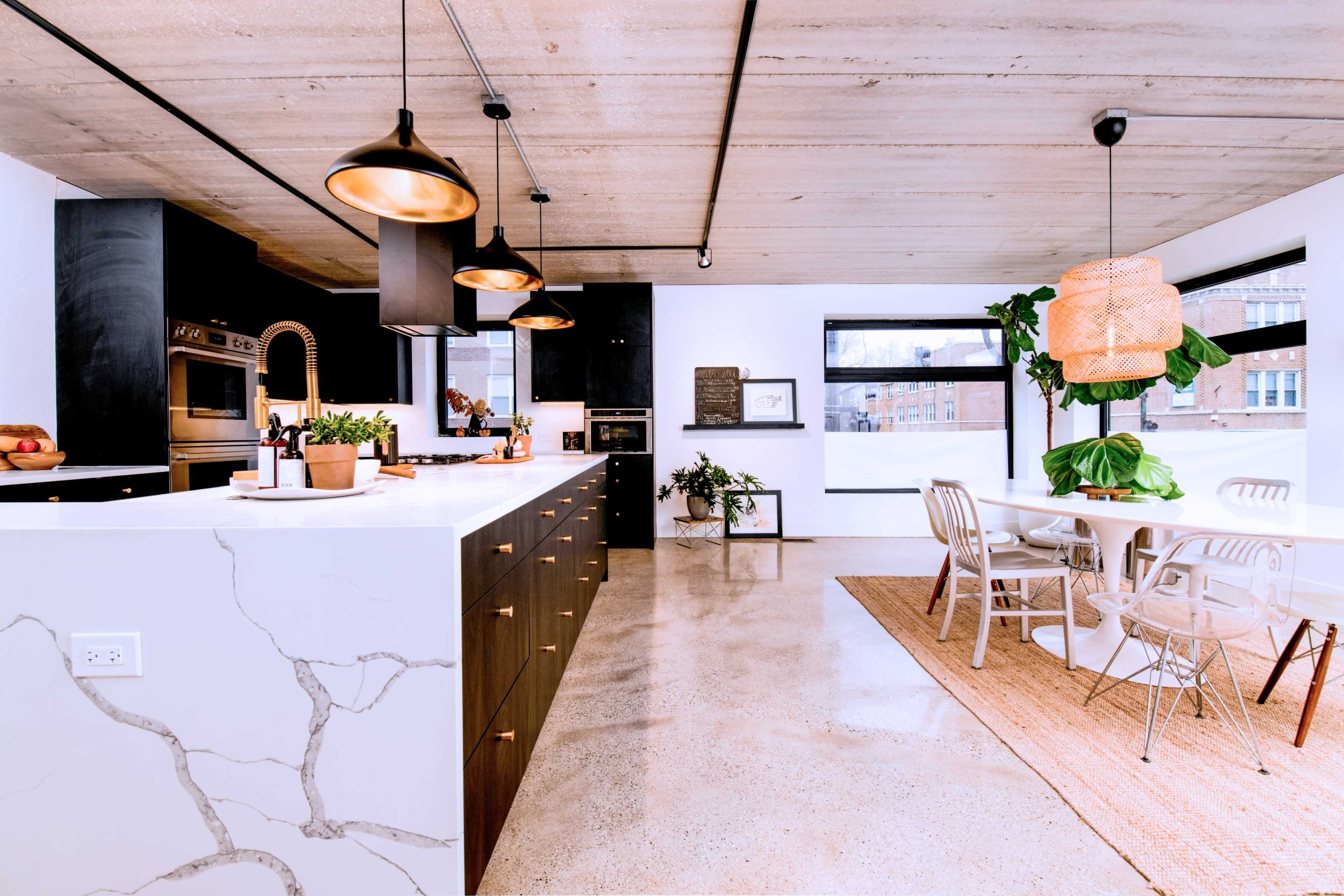 The image features a modern kitchen with a large island, dark cabinetry, stainless steel appliances, and a dining area with a round table and decorative plants.