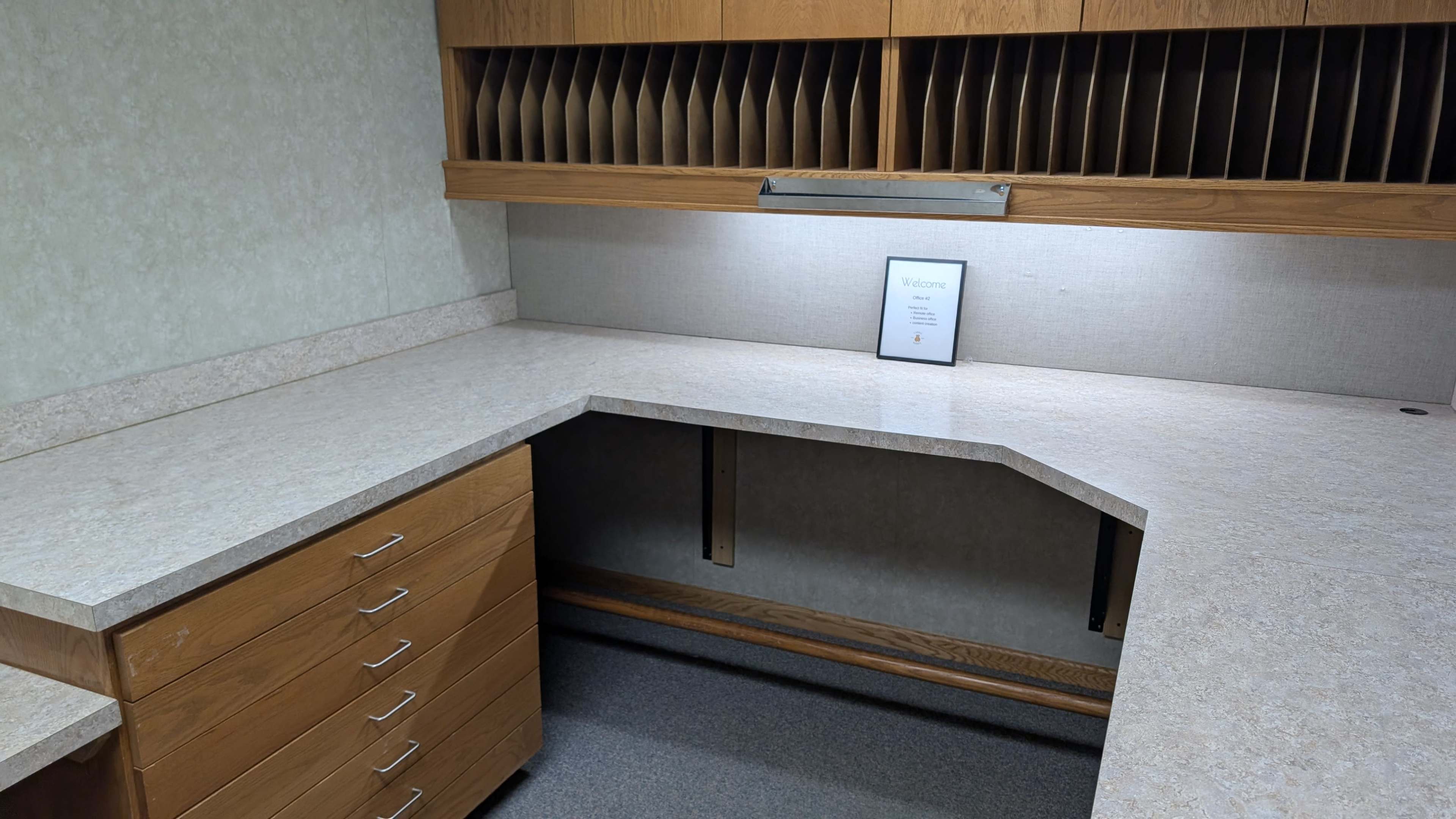 The image shows an empty office space featuring a corner desk with a light-colored countertop and wooden cabinets above containing vertical dividers.