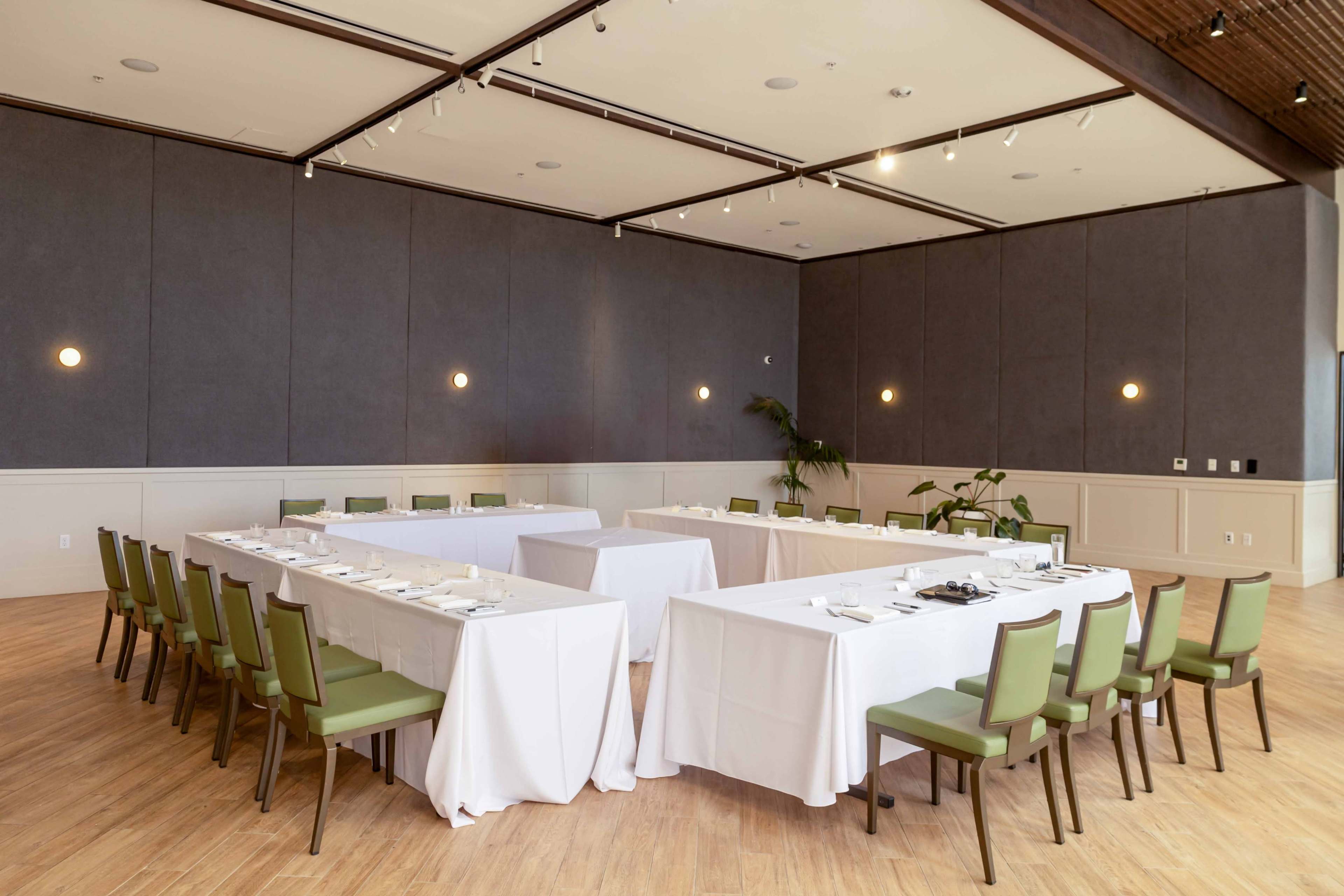 A spacious meeting room features two rectangular tables covered with white tablecloths arranged in a U-shape, surrounded by green chairs.