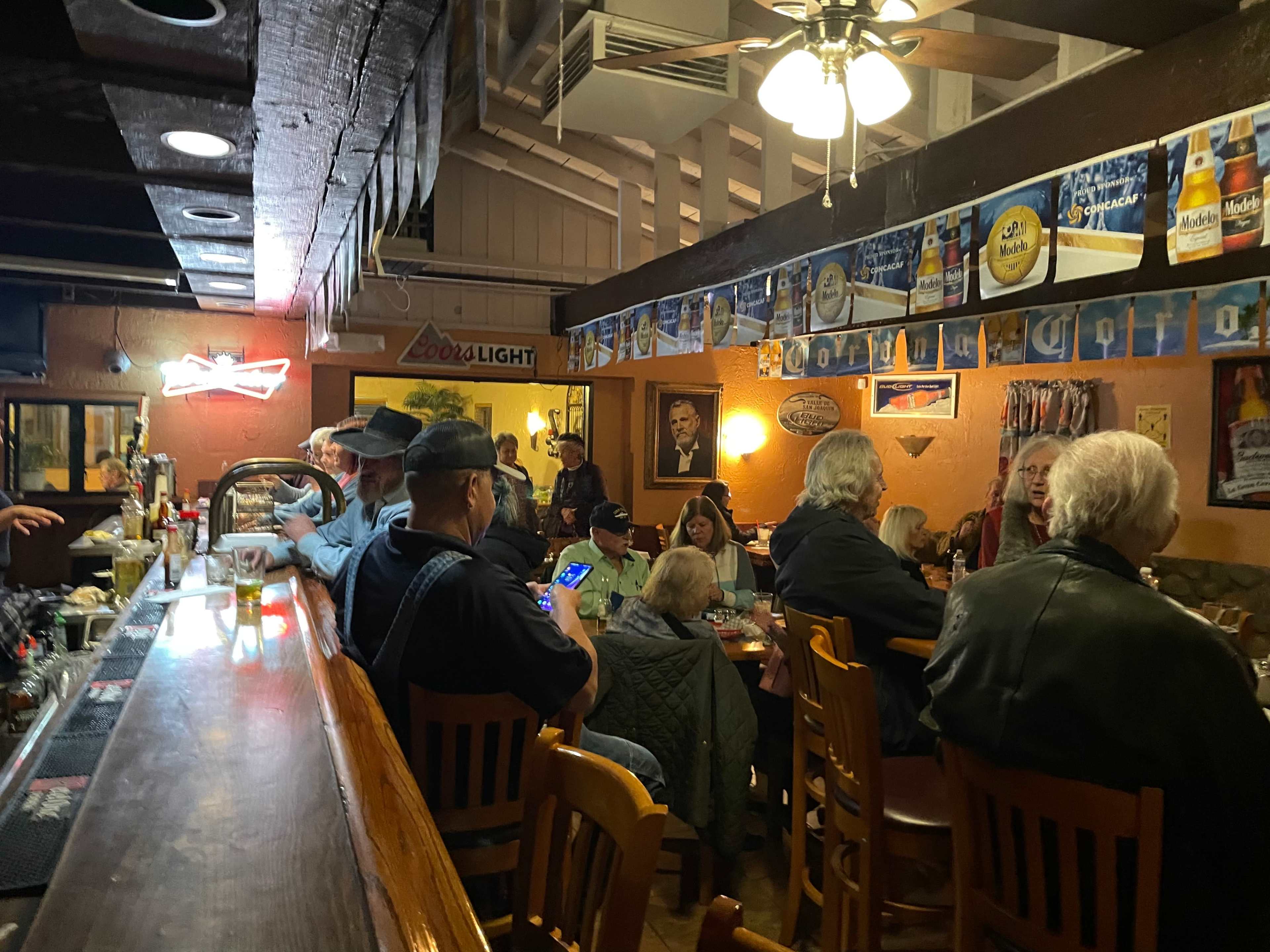 A crowded bar features patrons seated around wooden tables and a long bar, with beer advertisements and a portrait on the wall.