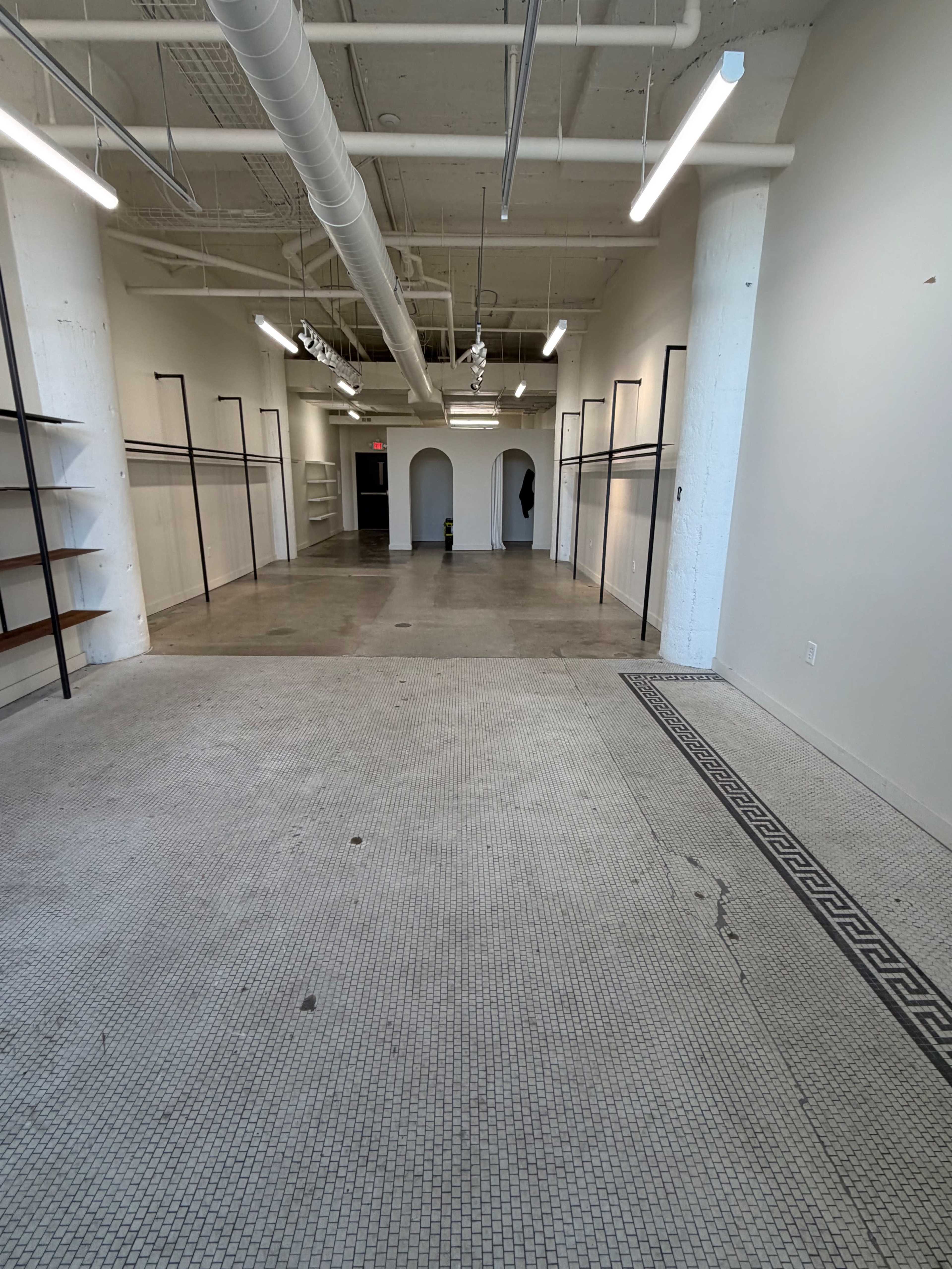 The image shows an empty commercial space with bare walls, a tiled floor, and shelving units along the sides.