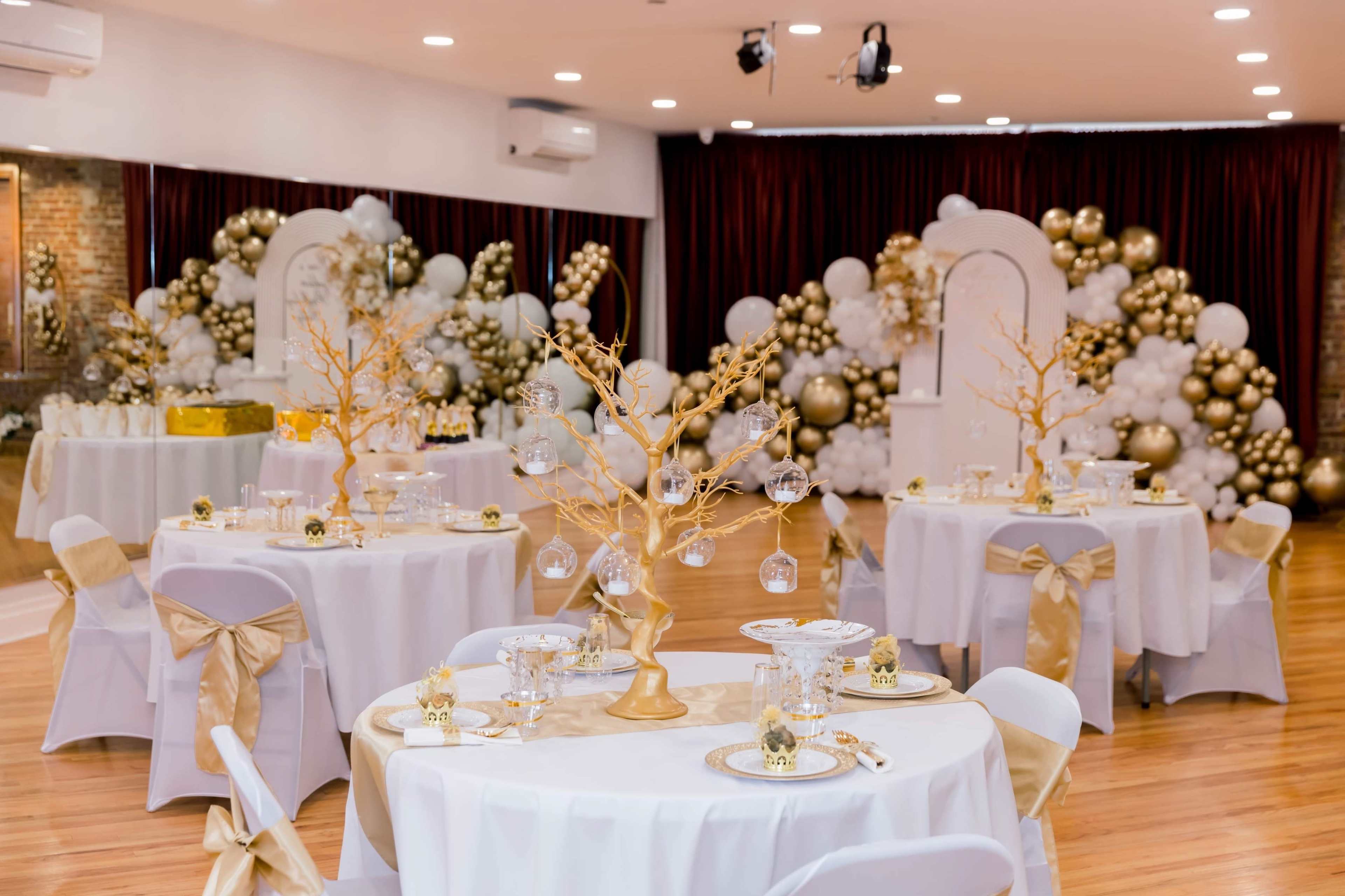 An elegantly decorated hall with tables set for an event, featuring white tablecloths and gold accents, surrounded by balloon decor in shades of gold and white.