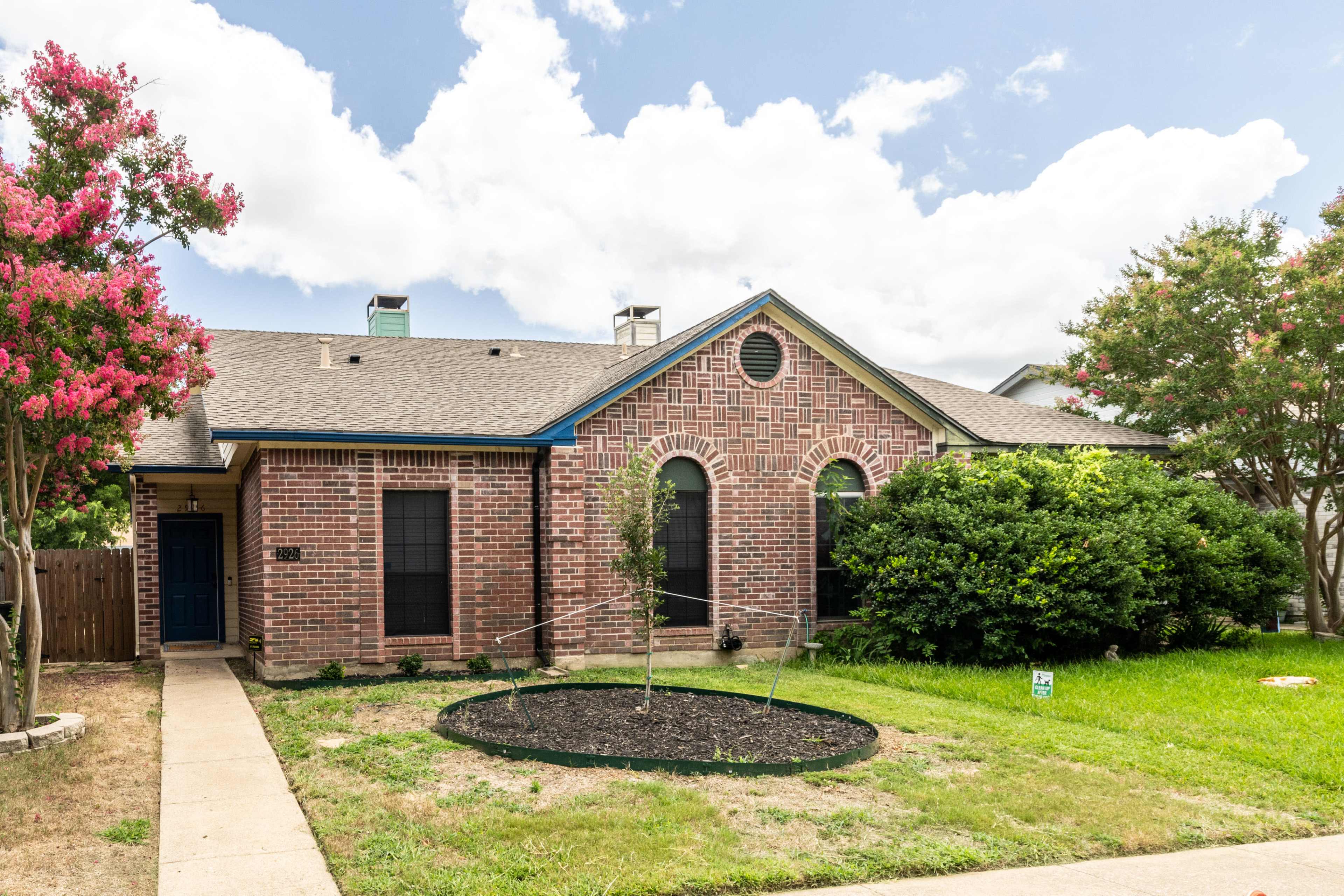 The image shows a single-story brick house with a landscaped yard, featuring a circular flower bed and flowering trees in the front.